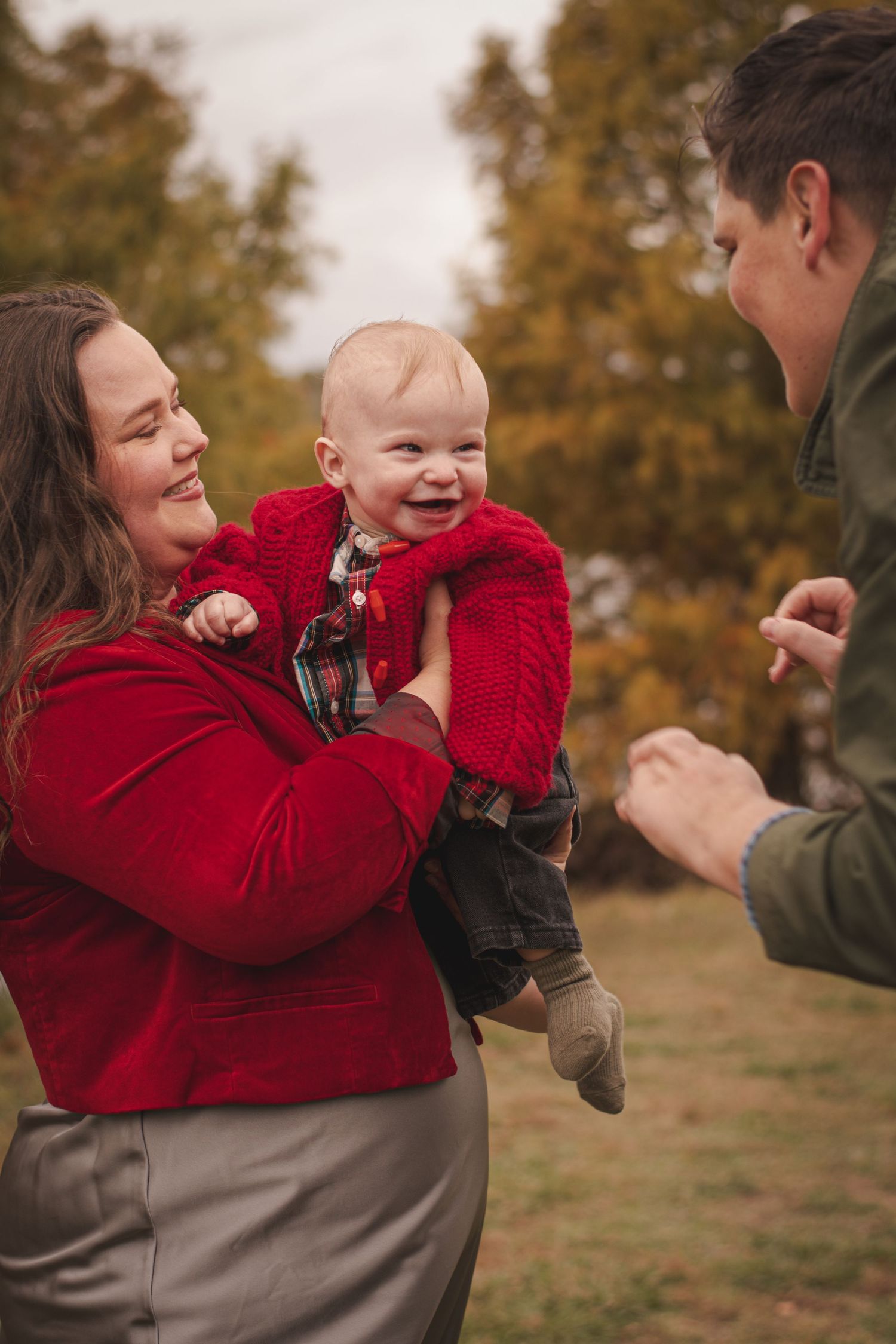 Parents smiling at laughing baby in red jacket during autumn outdoor family portrait.