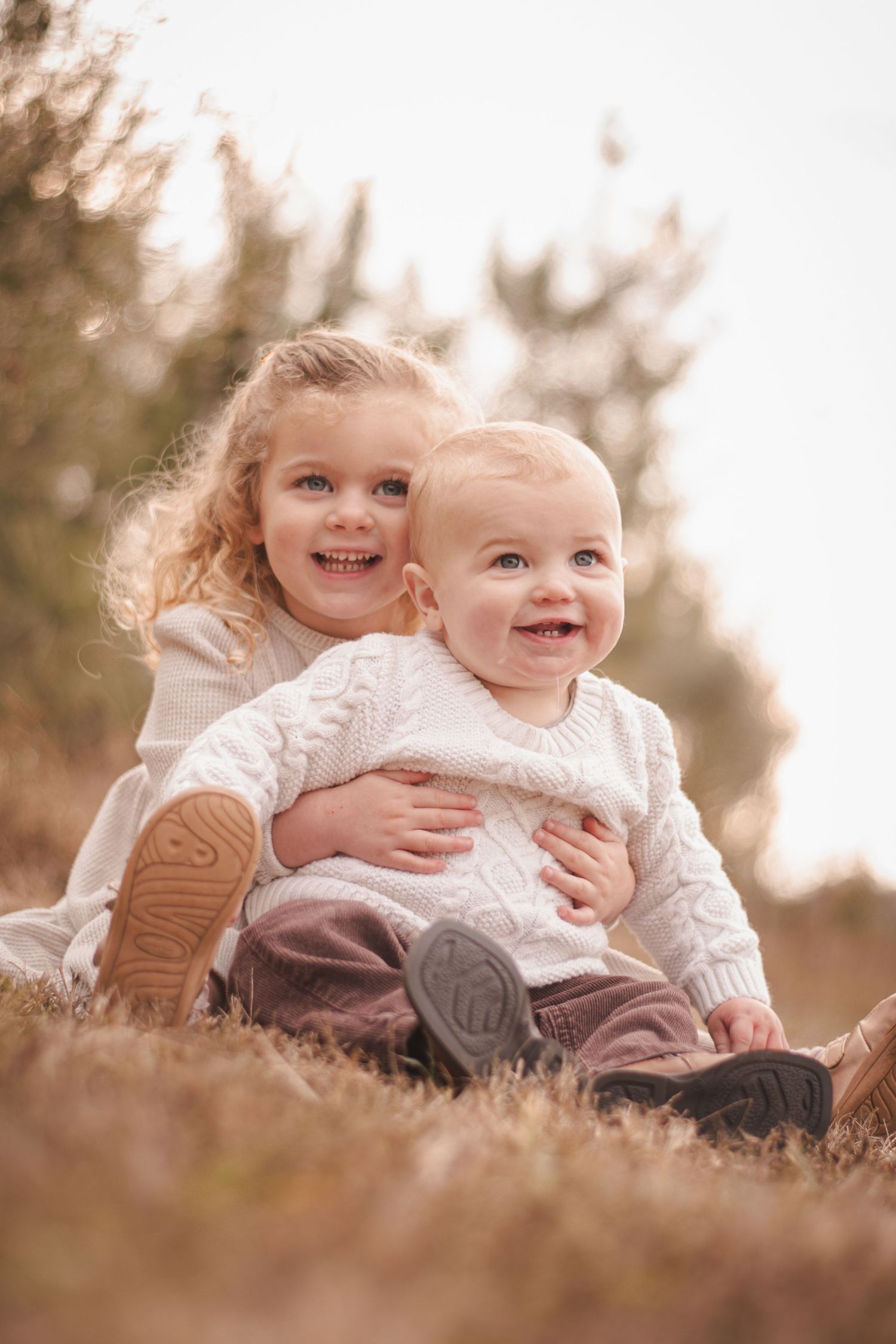 Two smiling children in white knit sweaters sit together outdoors in a golden field, older girl hugging baby sibling.