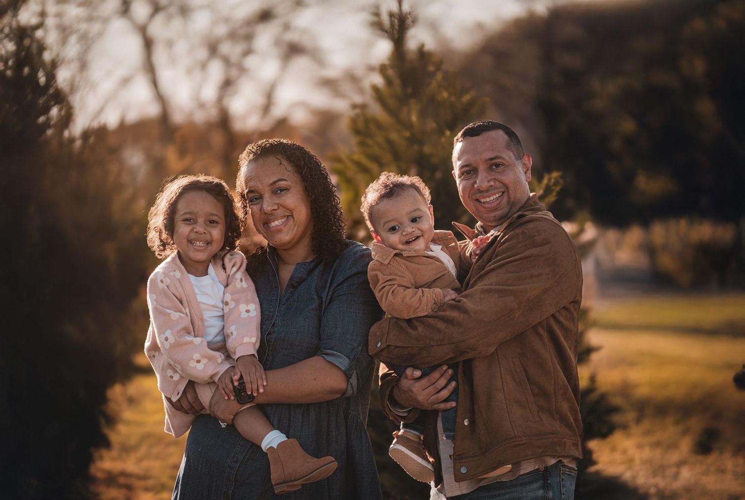 Smiling family of four posing outdoors at sunset with two young children in a festive setting.