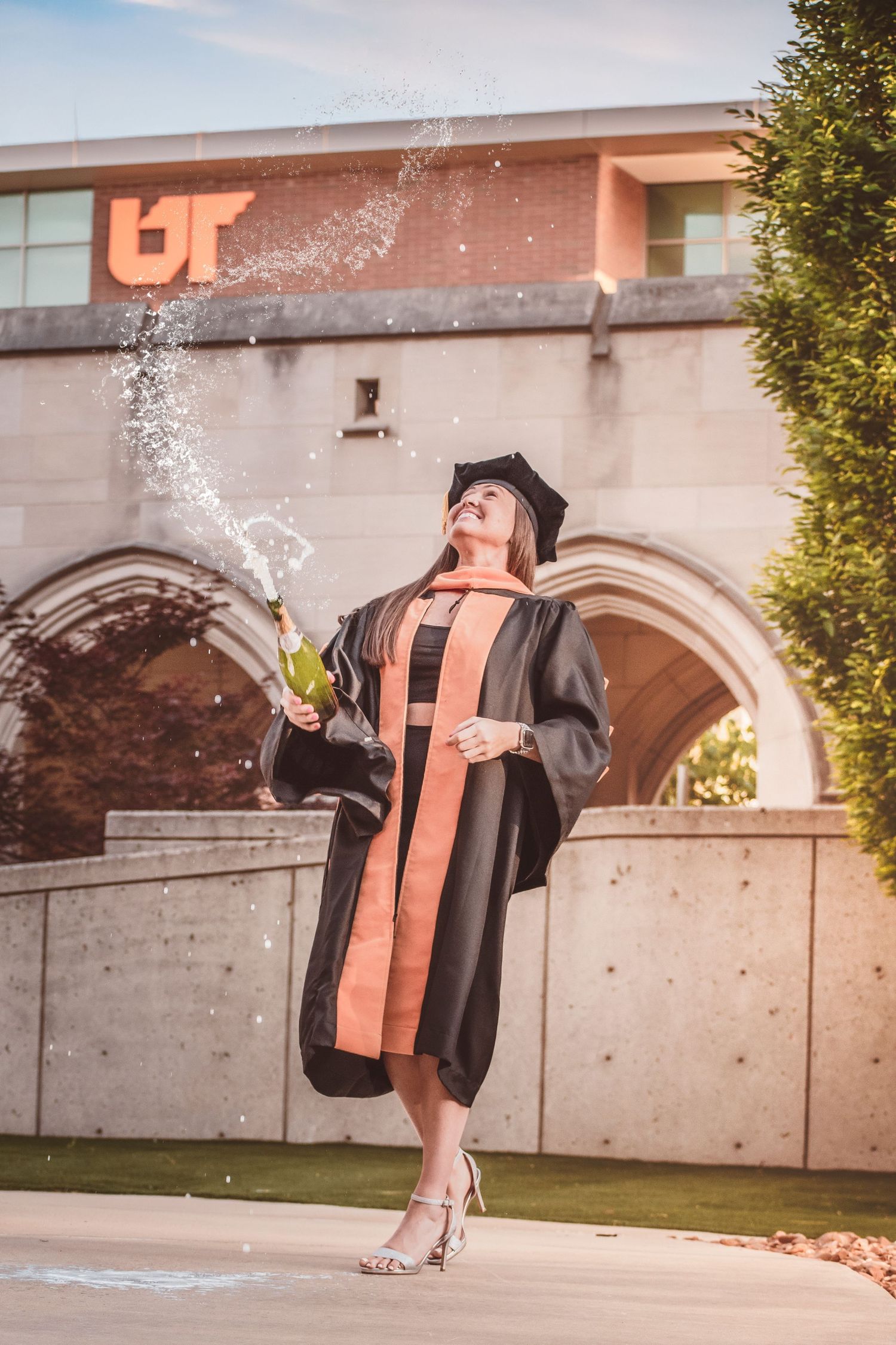 Graduate in black and orange regalia celebrates by popping champagne bottle outside UT building.