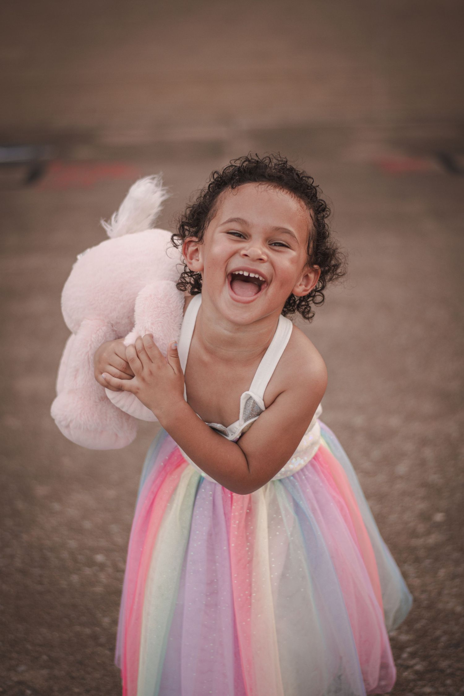 A joyful little girl in a rainbow tutu dress hugs a pink stuffed animal, laughing outdoors.