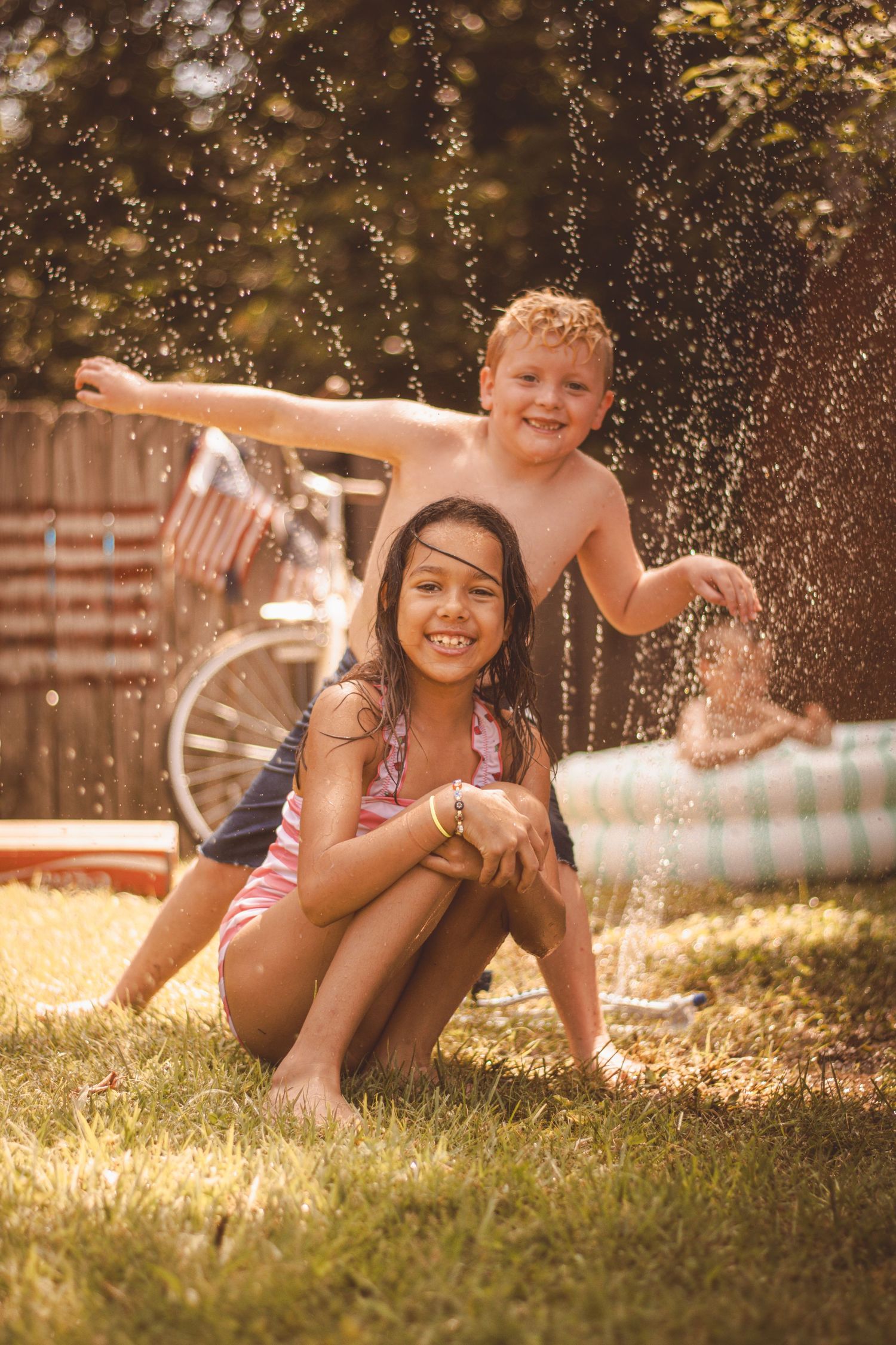 Two children playing in backyard sprinkler on sunny summer day, girl in swimsuit smiling while boy stands behind her.