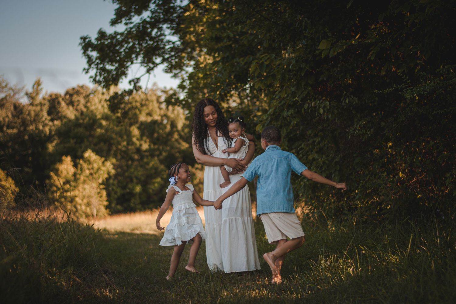 A mother in a white dress embraces her children outdoors in golden hour light near lush green trees. Lifestyle Photography
