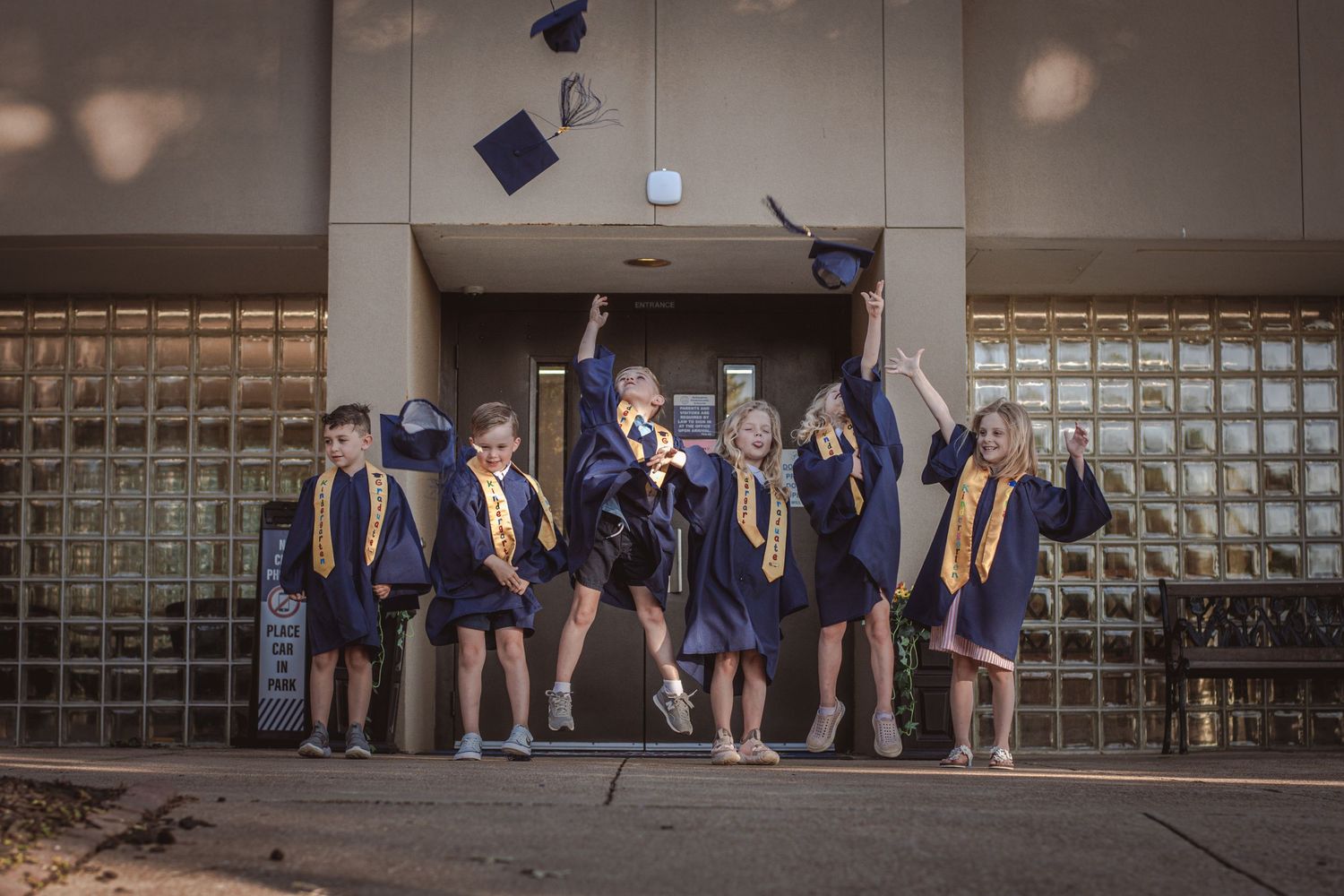 Six young children in blue graduation caps and gowns toss caps in the air outside school building.