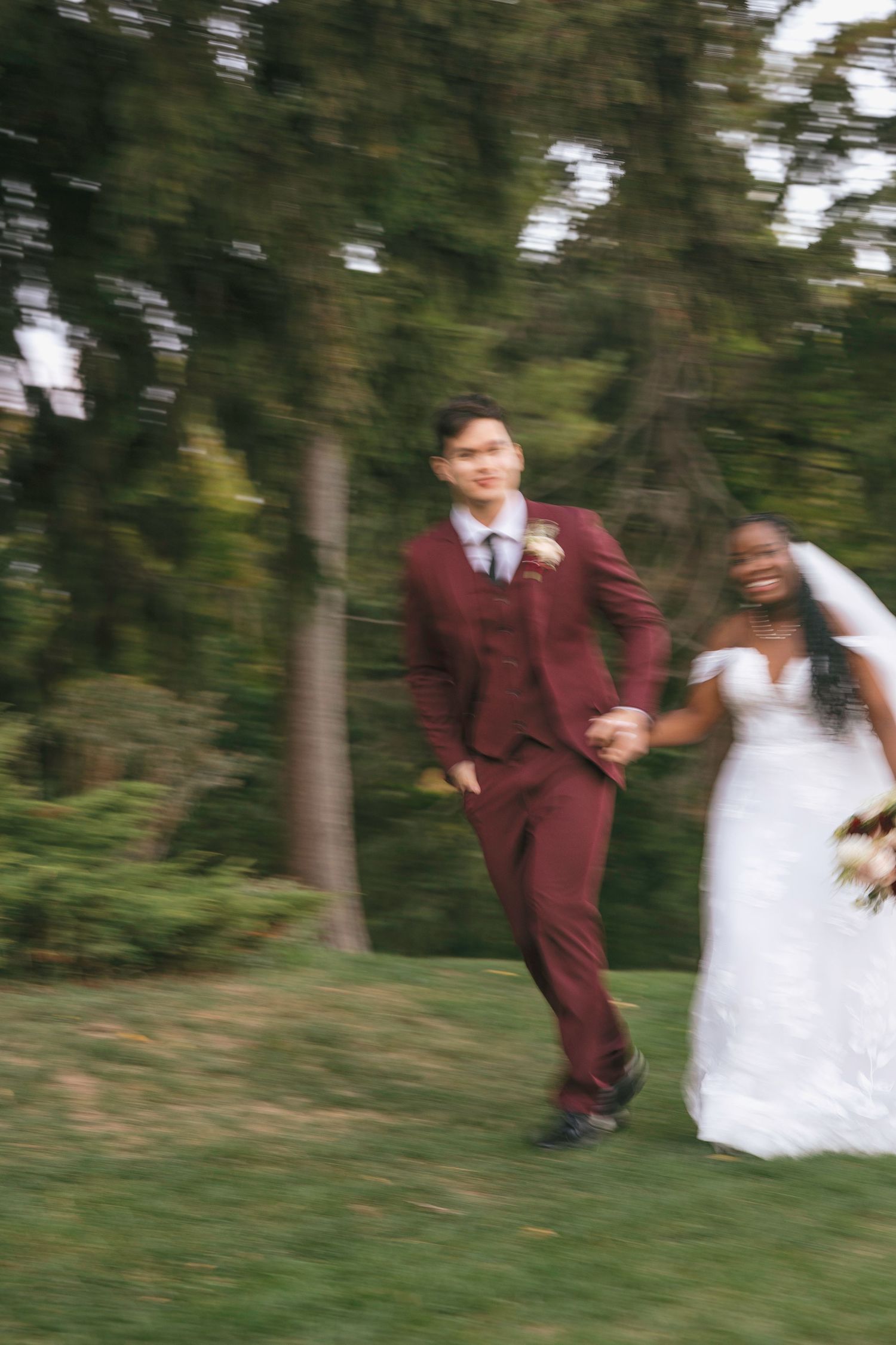 Blurred motion photo of interracial couple in wedding attire, groom in burgundy suit, bride in white gown, running outdoors.