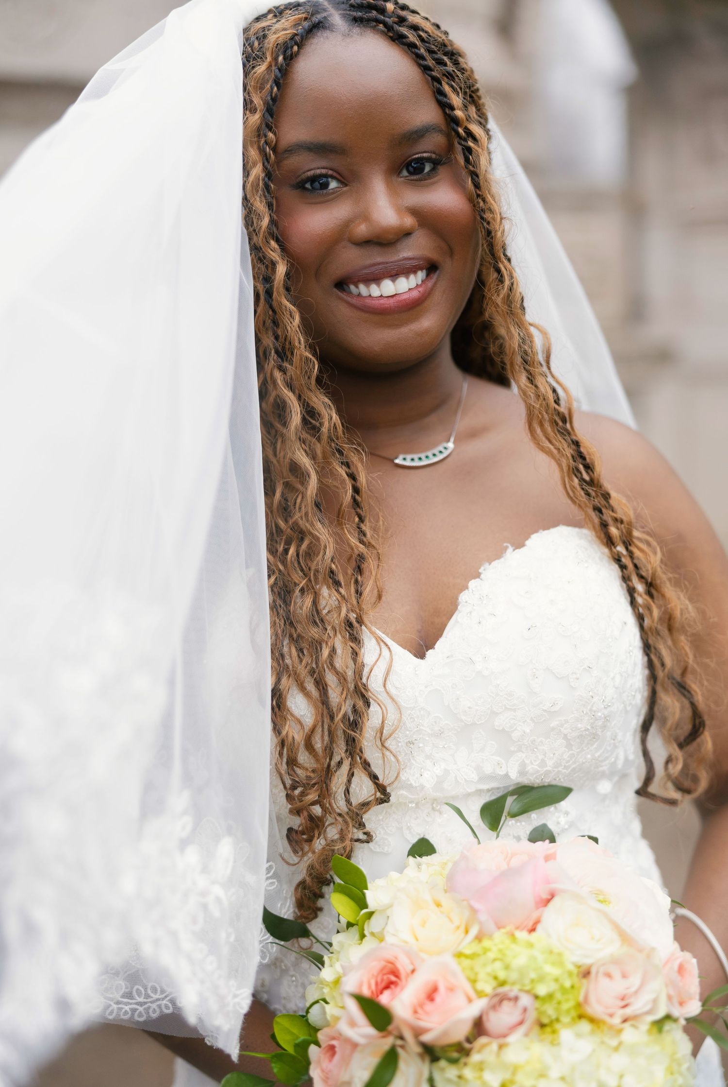 Smiling bride in white strapless lace gown and veil holds pink and white bouquet, with braided hair and diamond necklace.