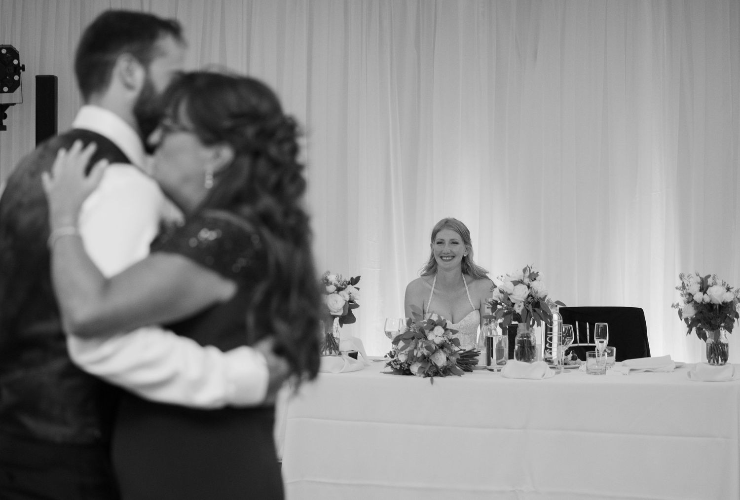 A couple shares a romantic dance at their wedding reception with white decor and floral arrangements in the background.