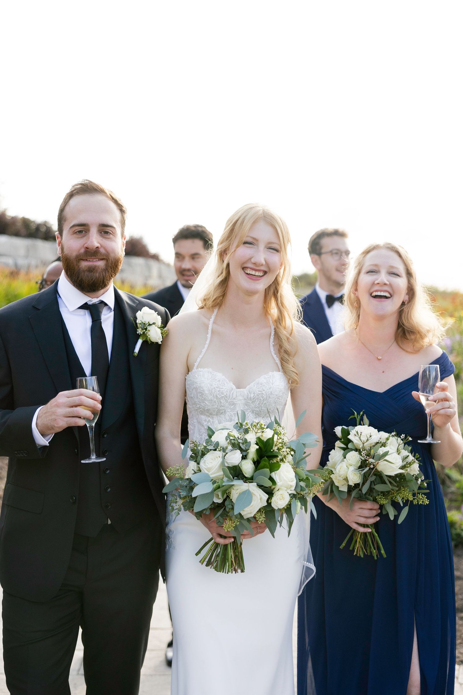 Wedding party in navy blue and white attire laughing together outdoors during golden hour celebration.