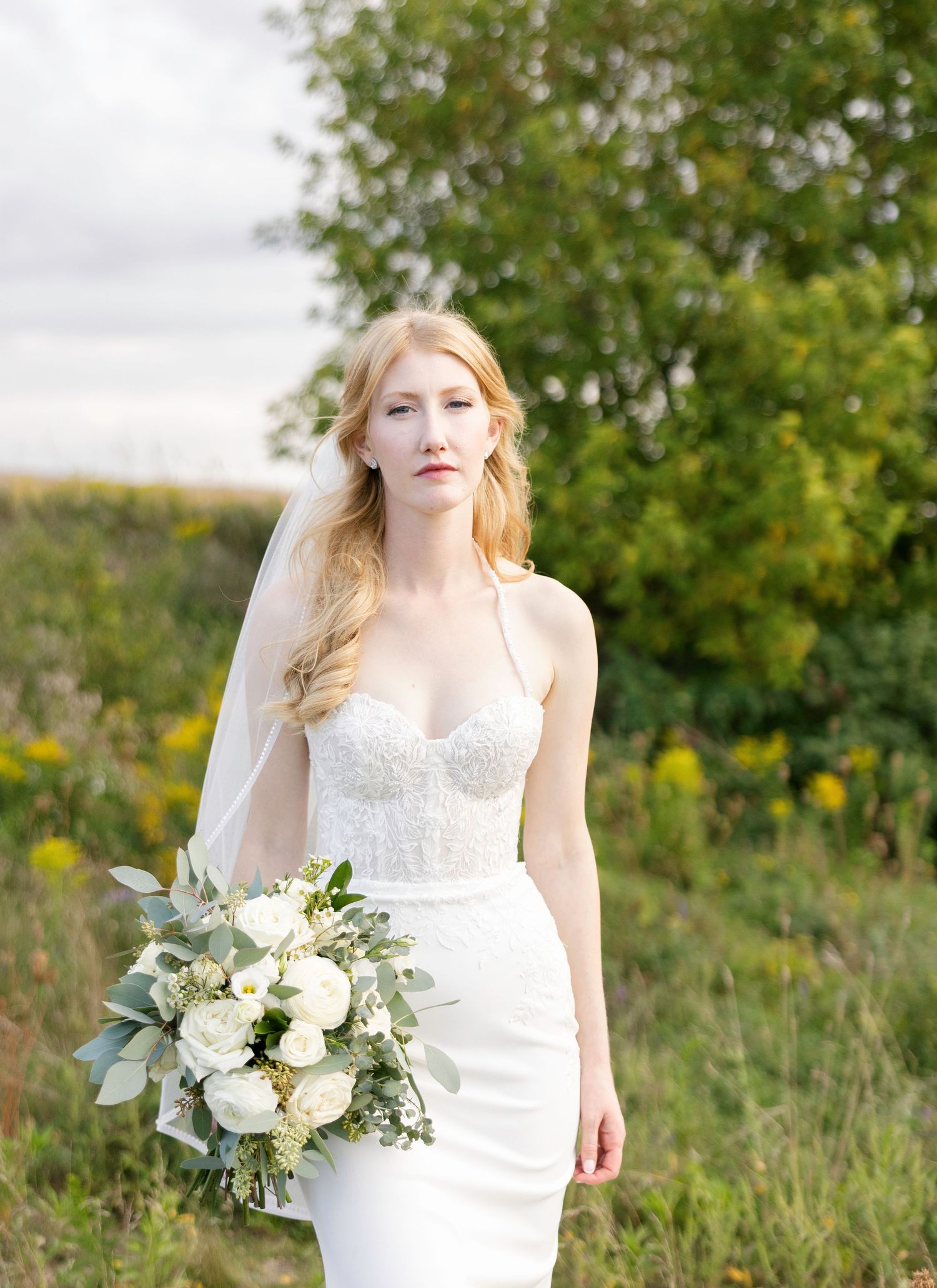 A bride in a white lace wedding dress holds a lush bouquet of white flowers and greenery in a natural outdoor setting.