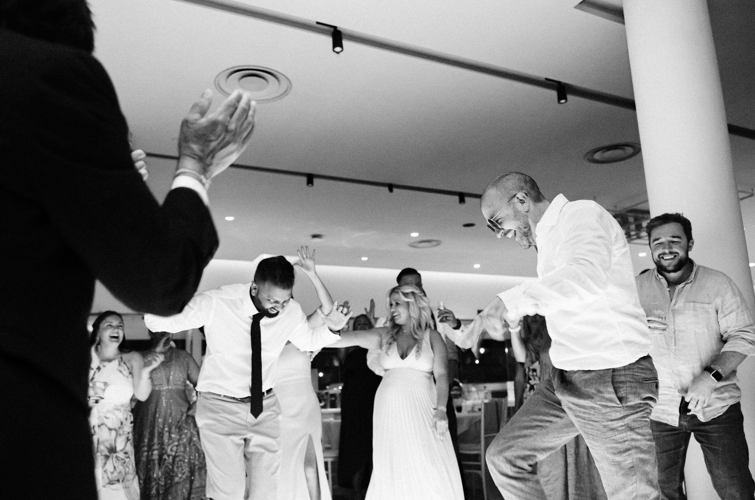 Black and white photograph of people dancing and celebrating at a wedding reception in an indoor venue.