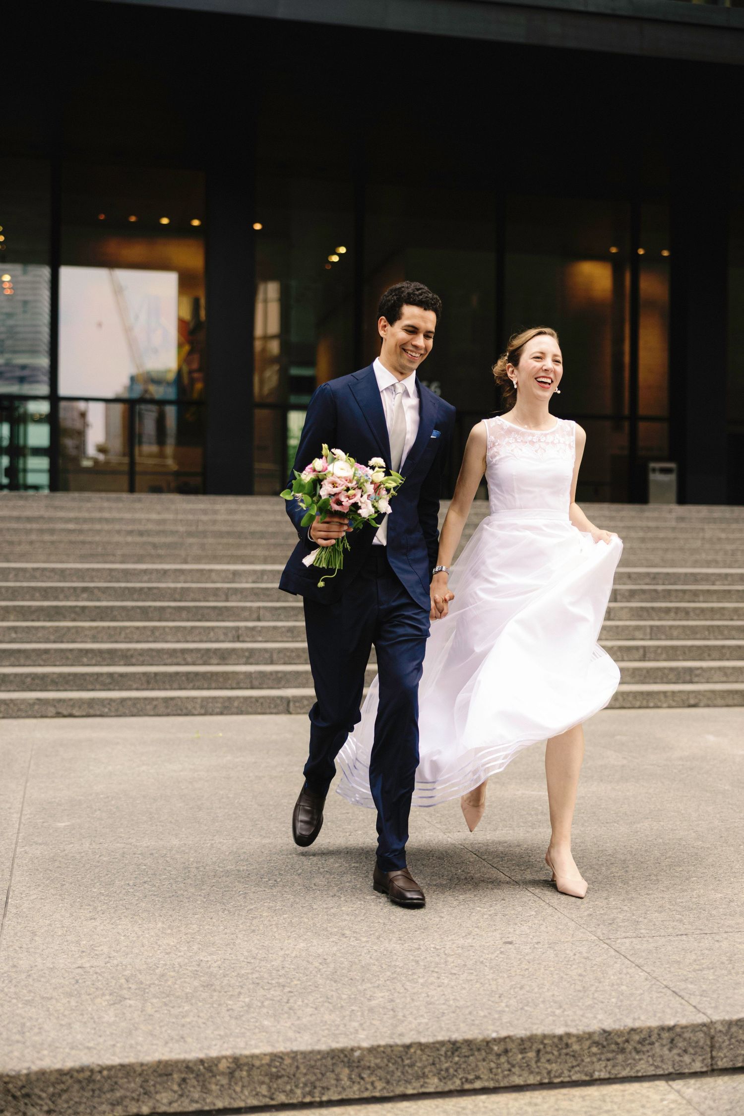 Couple in wedding attire walking and laughing on concrete steps outside building.