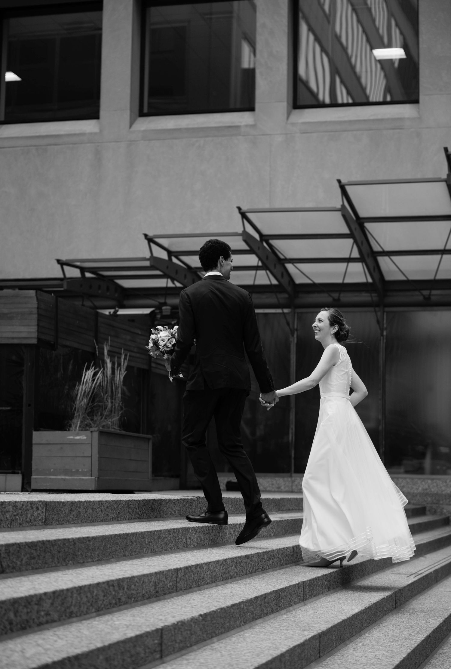 A wedding couple walks down a modern building's concrete steps in black and white.
