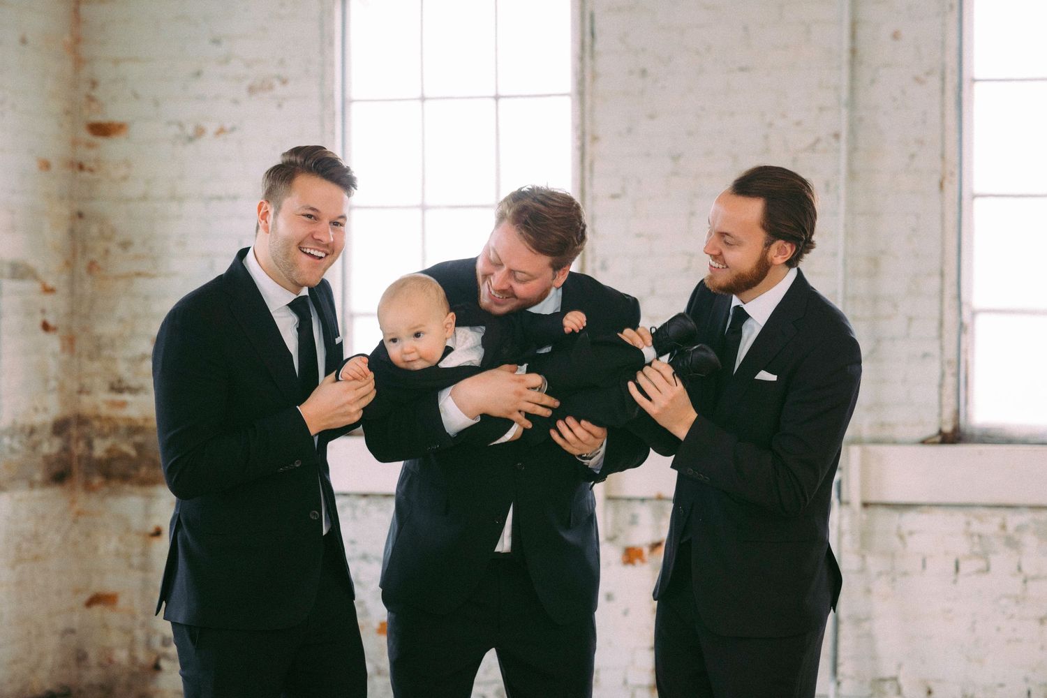 Groomsmen in formal attire share candid laughs while sitting on wooden floor.