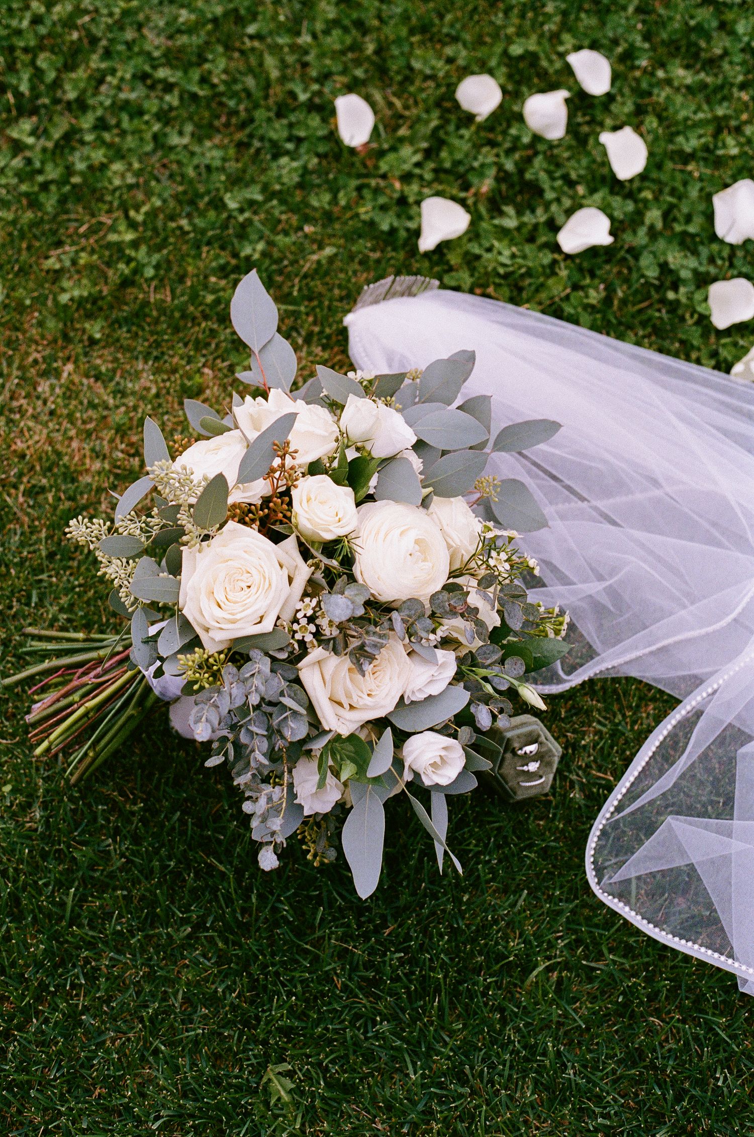 A delicate bridal bouquet with white roses and eucalyptus leaves lies on grass next to scattered petals and a sheer veil.