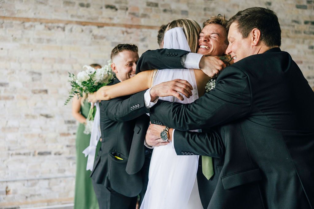 Groomsmen in black suits share an emotional group hug at a wedding celebration against a rustic brick wall.