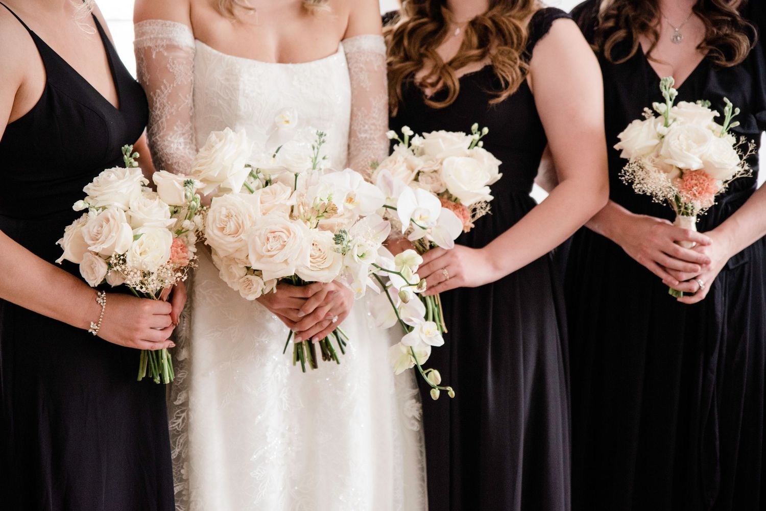 Wedding party members holding white floral bouquets lined up in black dresses.
