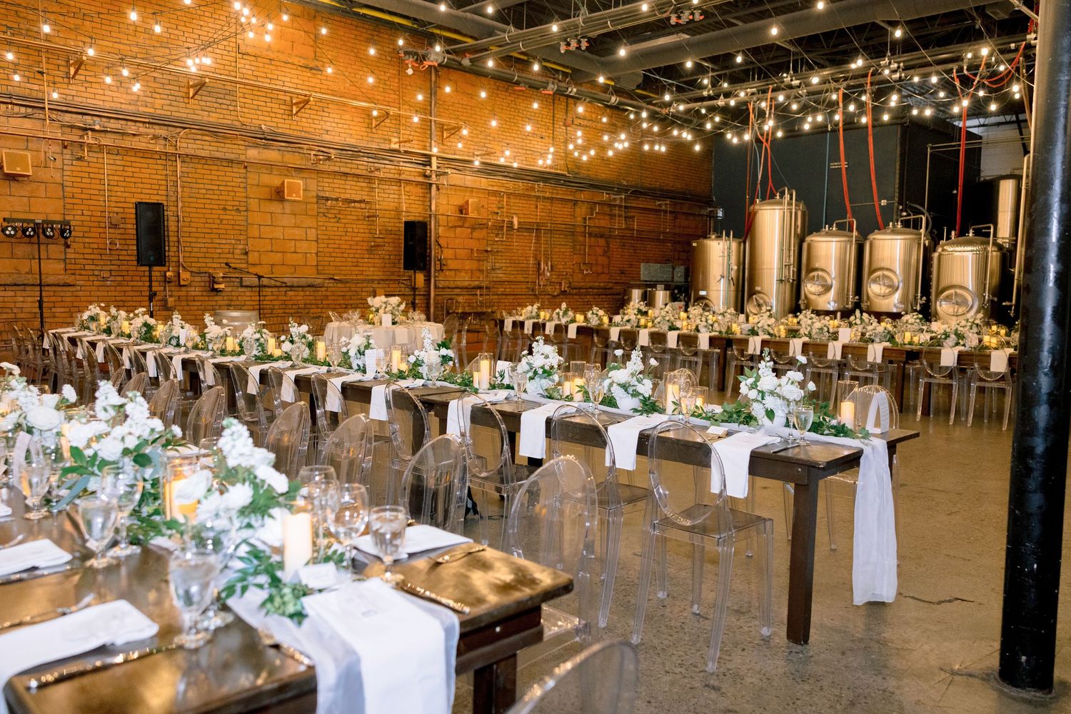 Elegant wedding reception in a brewery venue with string lights, ghost chairs, and lush greenery centerpieces on rustic tables.