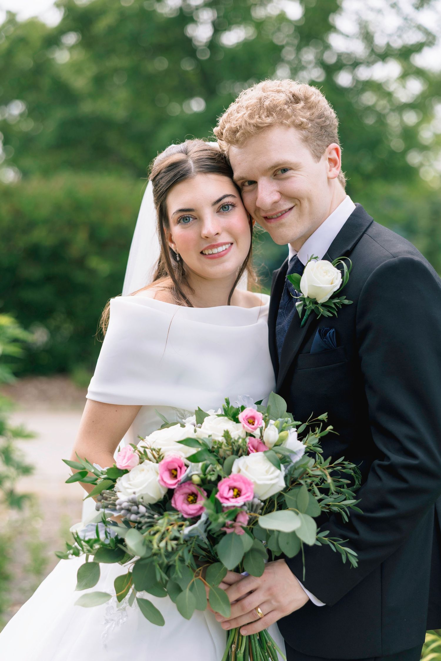 Smiling bride and groom pose outdoors holding a pink and white floral bouquet, bride in white gown with veil, groom in dark suit.
