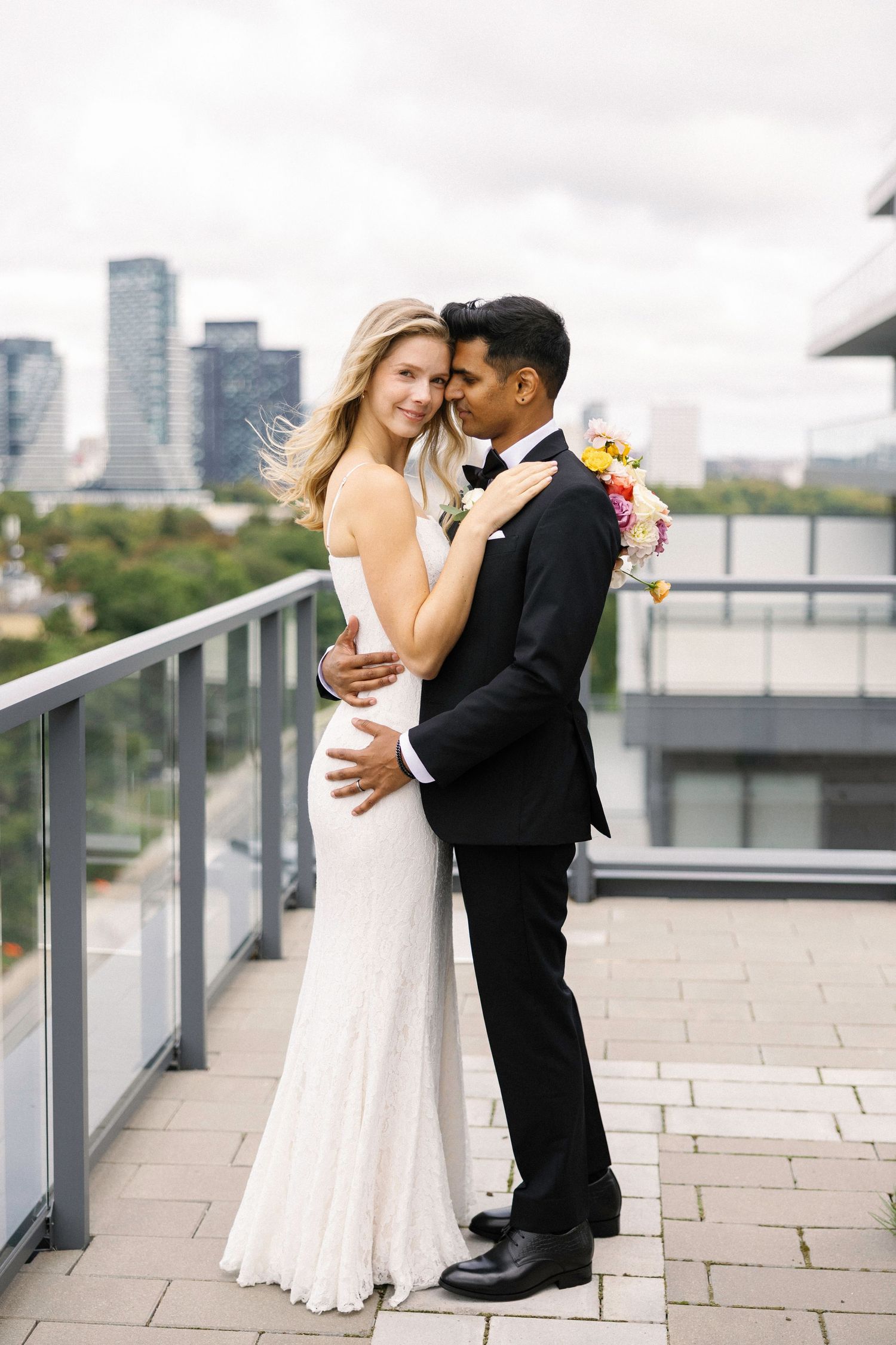 Bride and groom embrace on rooftop terrace with city skyline backdrop, bride in white gown, groom in black tuxedo.