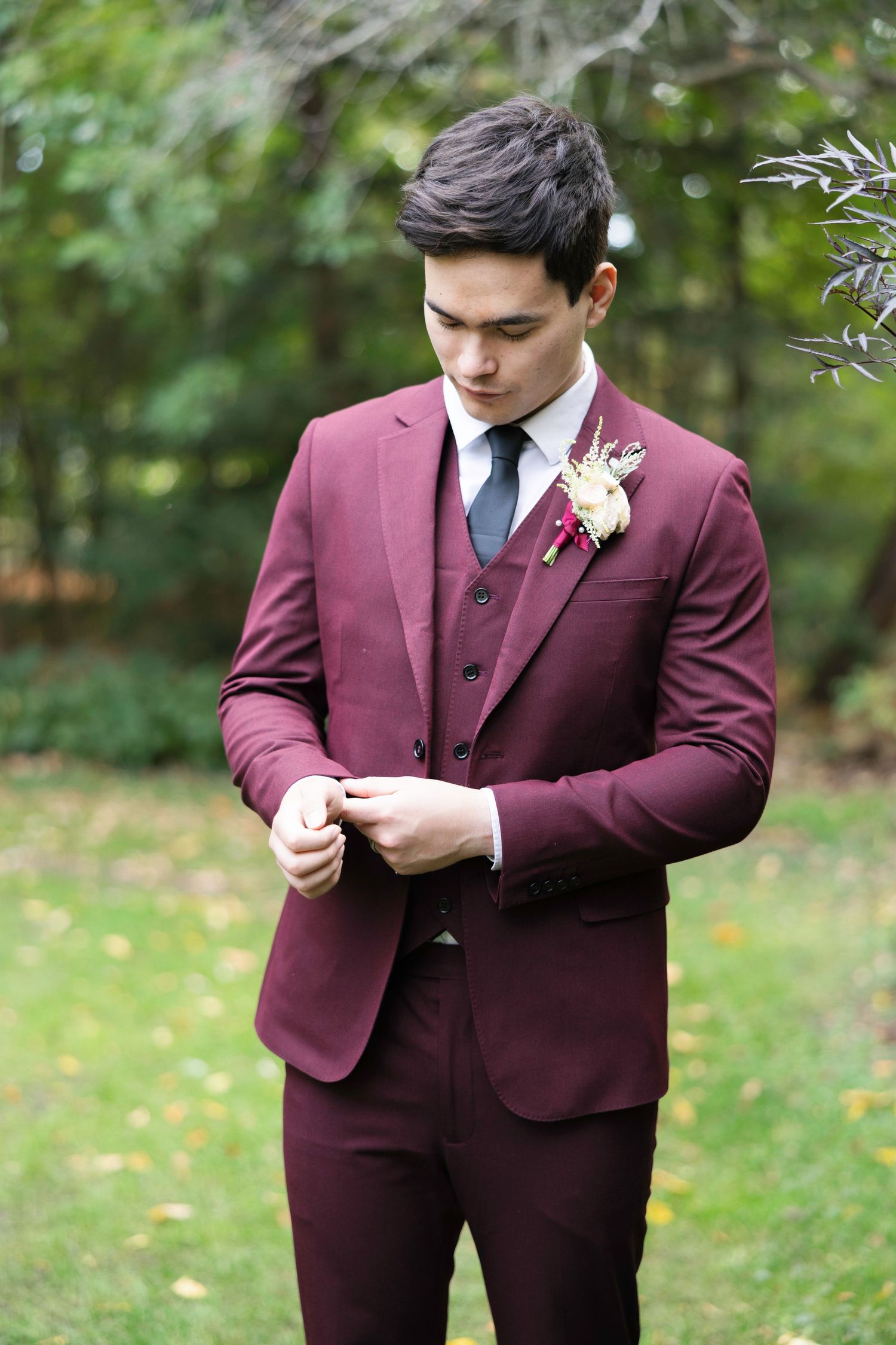 A person in a burgundy suit adjusts their boutonniere while standing outdoors in a garden setting.