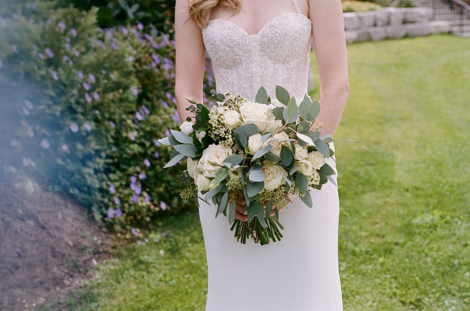 A bridal bouquet of white roses and eucalyptus leaves held against a white wedding dress in an outdoor garden setting.