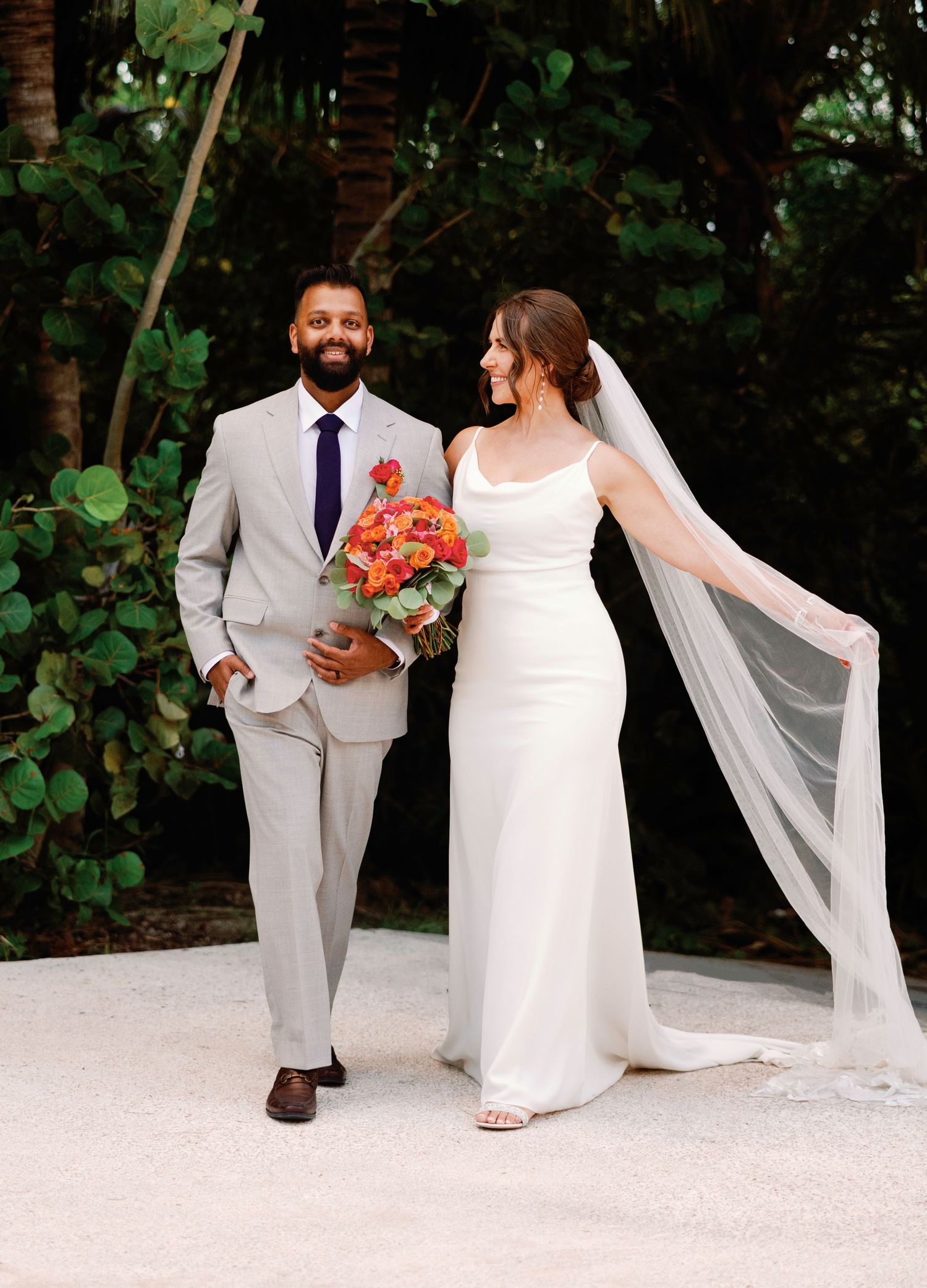 A couple in wedding attire shares intimate moments during their outdoor garden ceremony with flowing veil and coral bouquet.