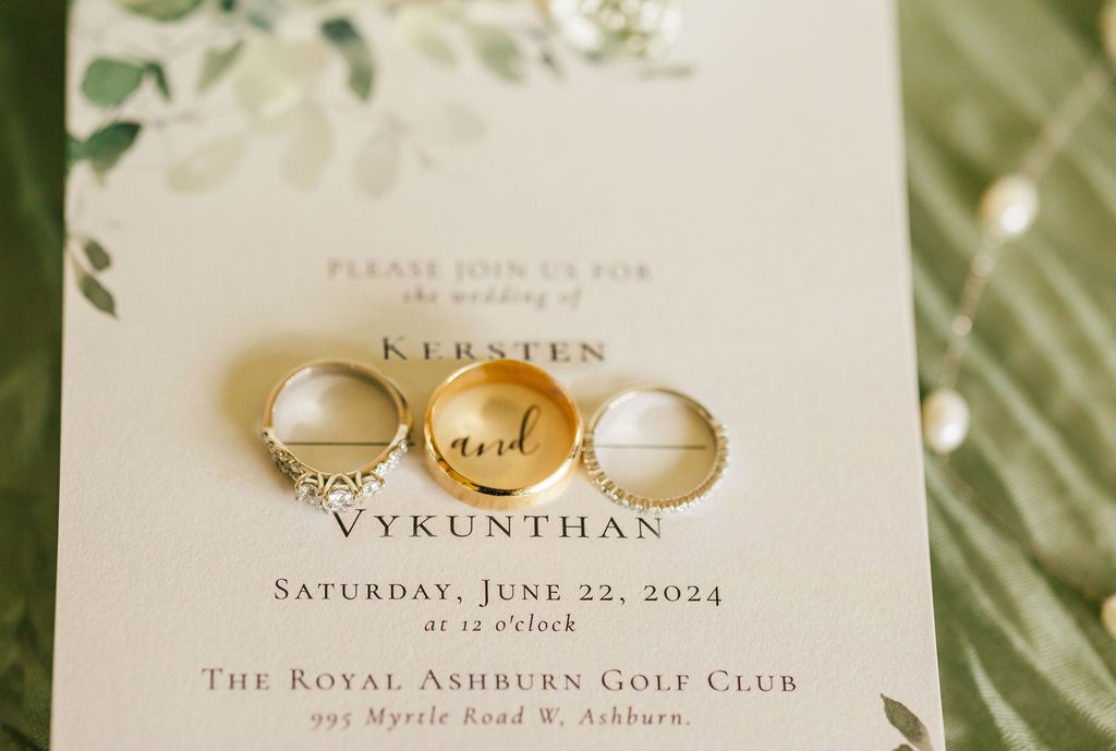 Elegant wedding invitation and gold rings displayed on light green textured background.