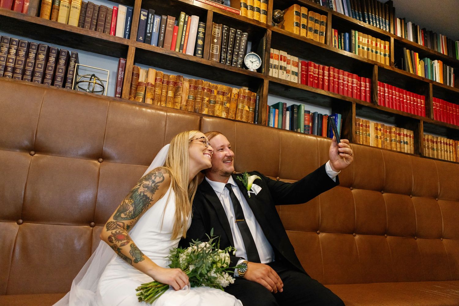 A couple takes a selfie while sitting on a brown leather couch in a library with vintage books on shelves behind them.