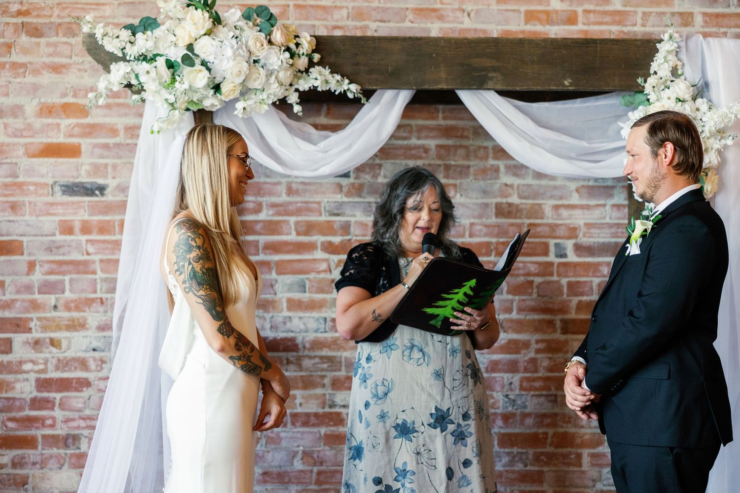 Wedding ceremony in progress under floral arch with exposed brick backdrop.