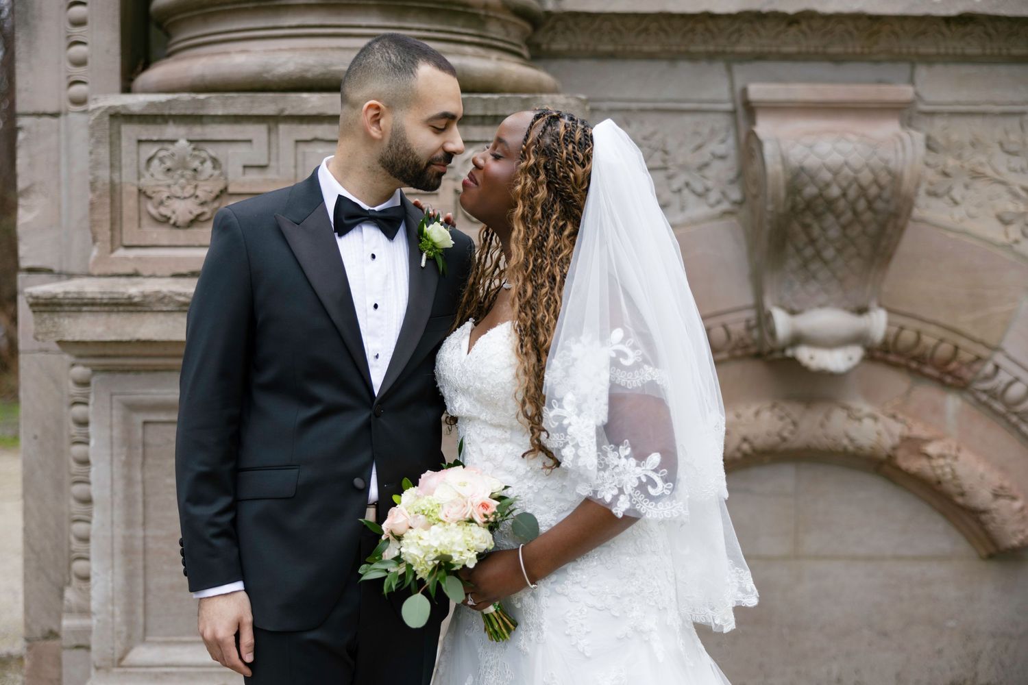 Bride and groom share a romantic moment outdoors, she in lace wedding gown with veil, he in black tuxedo.
