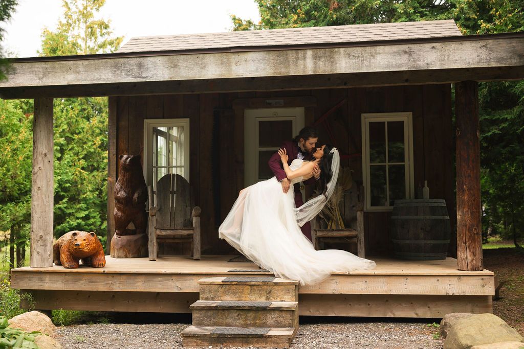 Bride and groom share romantic embrace on rustic wooden cabin porch surrounded by lush greenery.