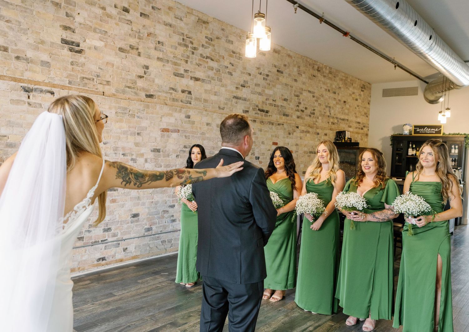 A panoramic view of a bridal party getting ready in a rustic brick-walled room with green dresses hanging on racks.