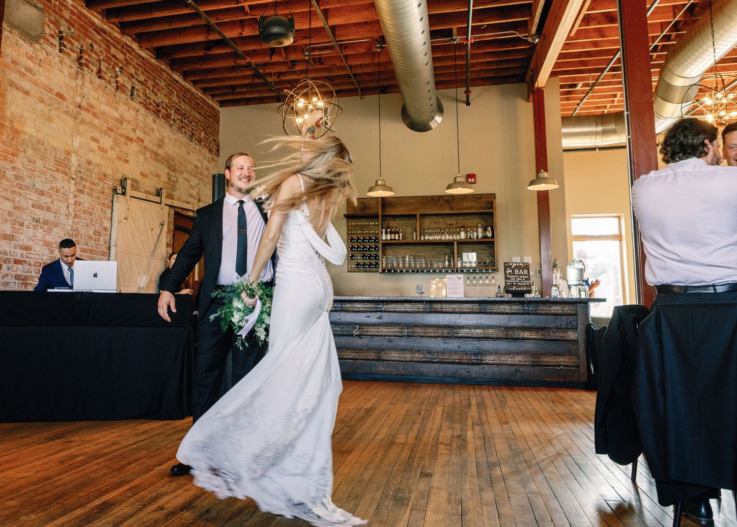 A wedding reception venue with exposed brick walls and rustic wooden floors features multiple candid dance moments.