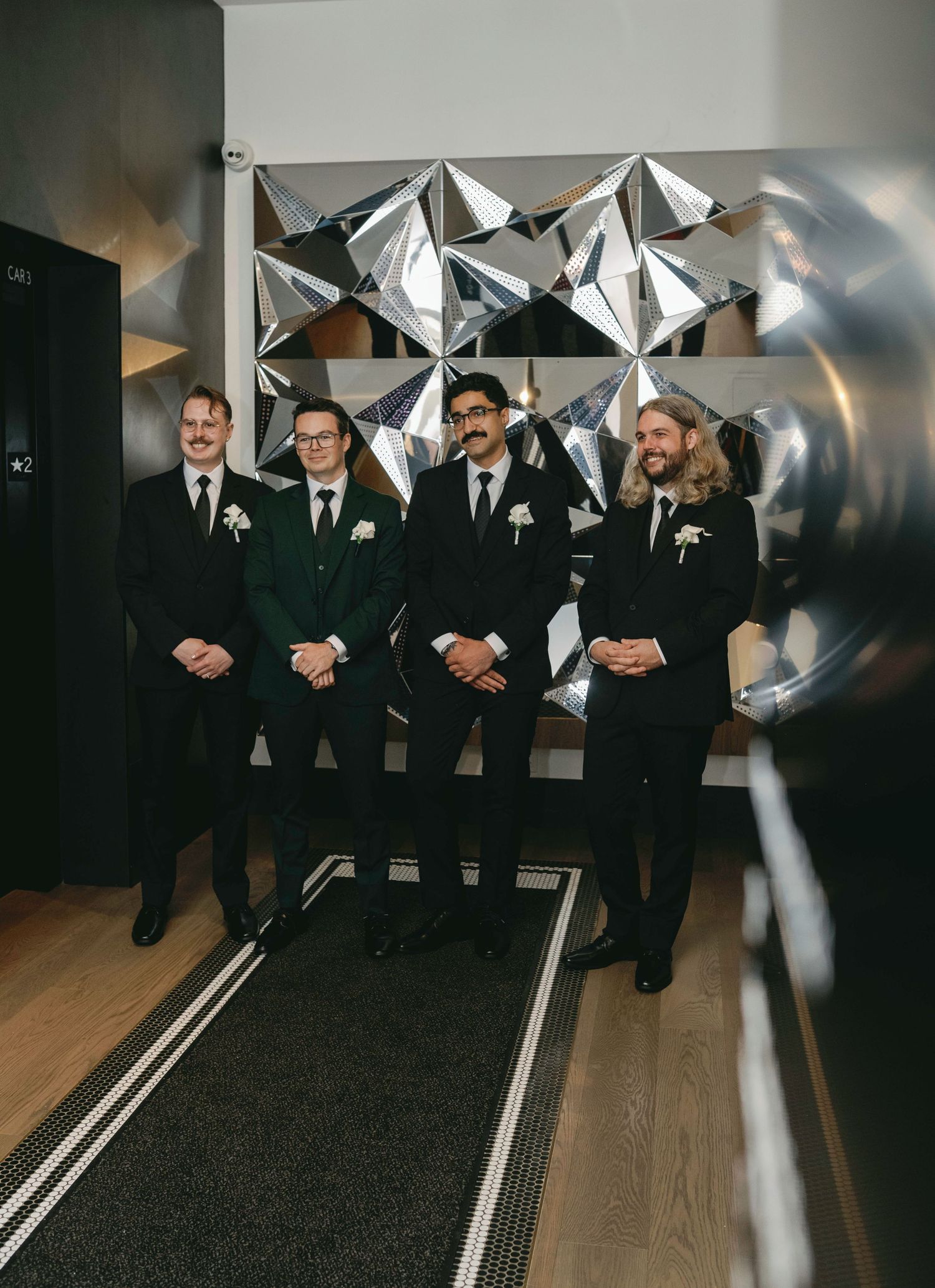 Wedding party members in black suits pose in front of a geometric silver wall decoration.