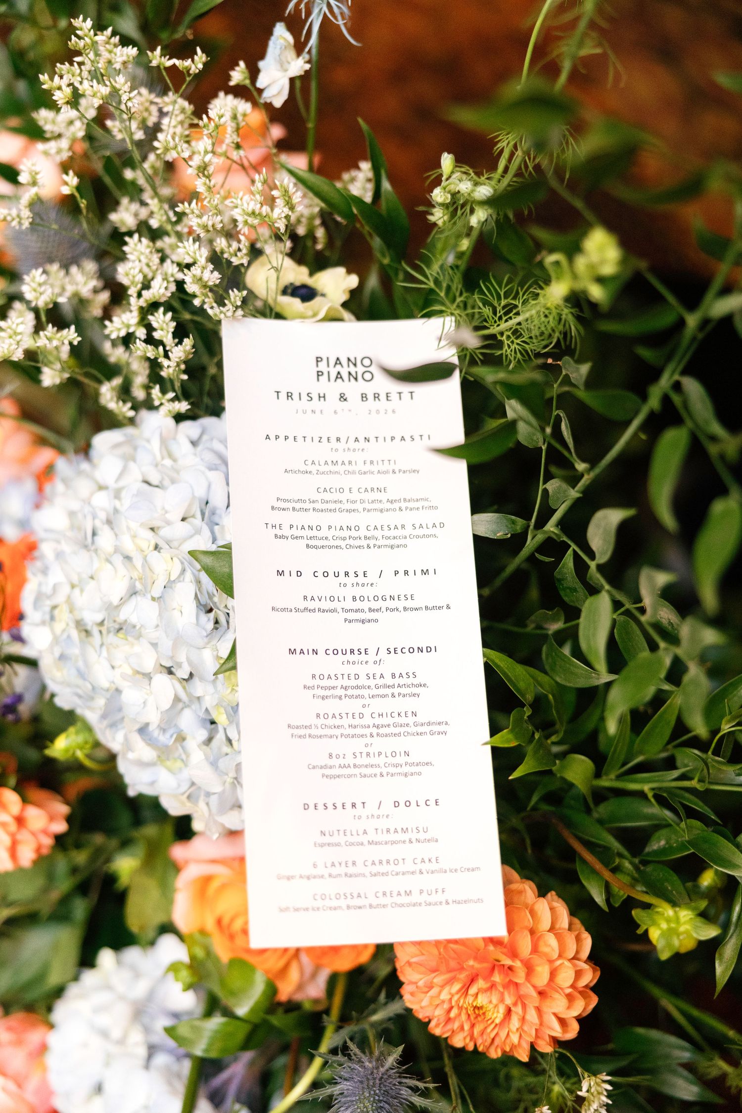 Elegant wedding menu card displayed among lush floral arrangements with hydrangeas and orange blooms.