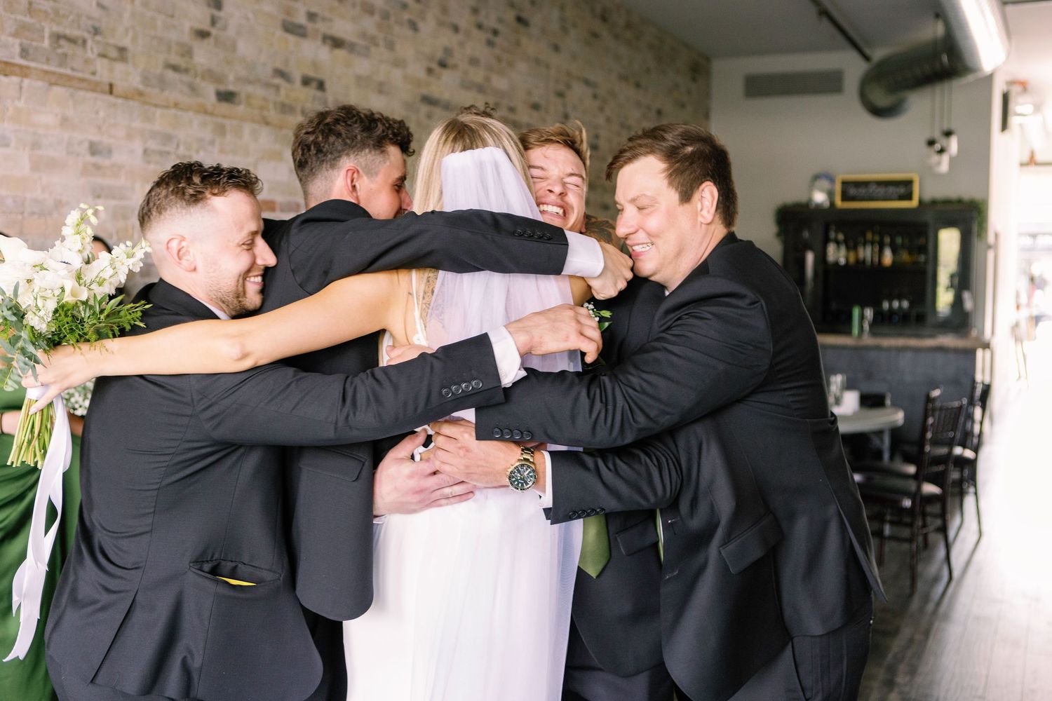 Wedding party embraces in a group hug while laughing and celebrating in an indoor venue.