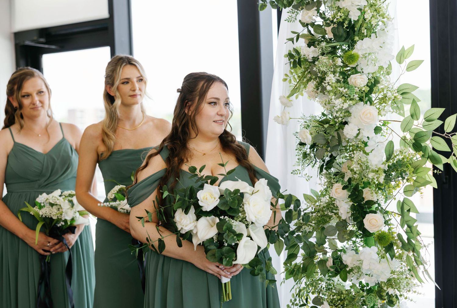 Bridesmaids in sage green dresses holding white bouquets pose together at wedding venue.