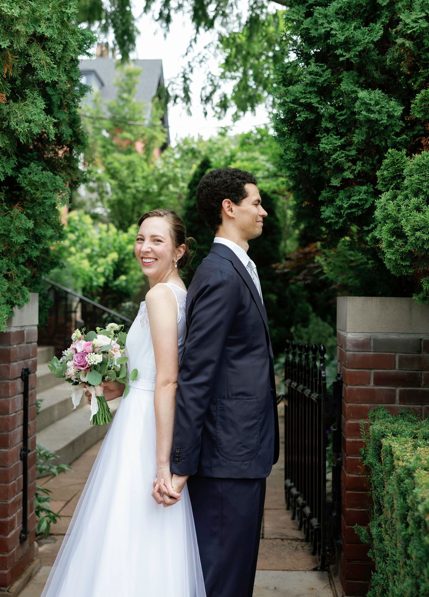 Couple walking along brick pathway surrounded by lush greenery in wedding attire.
