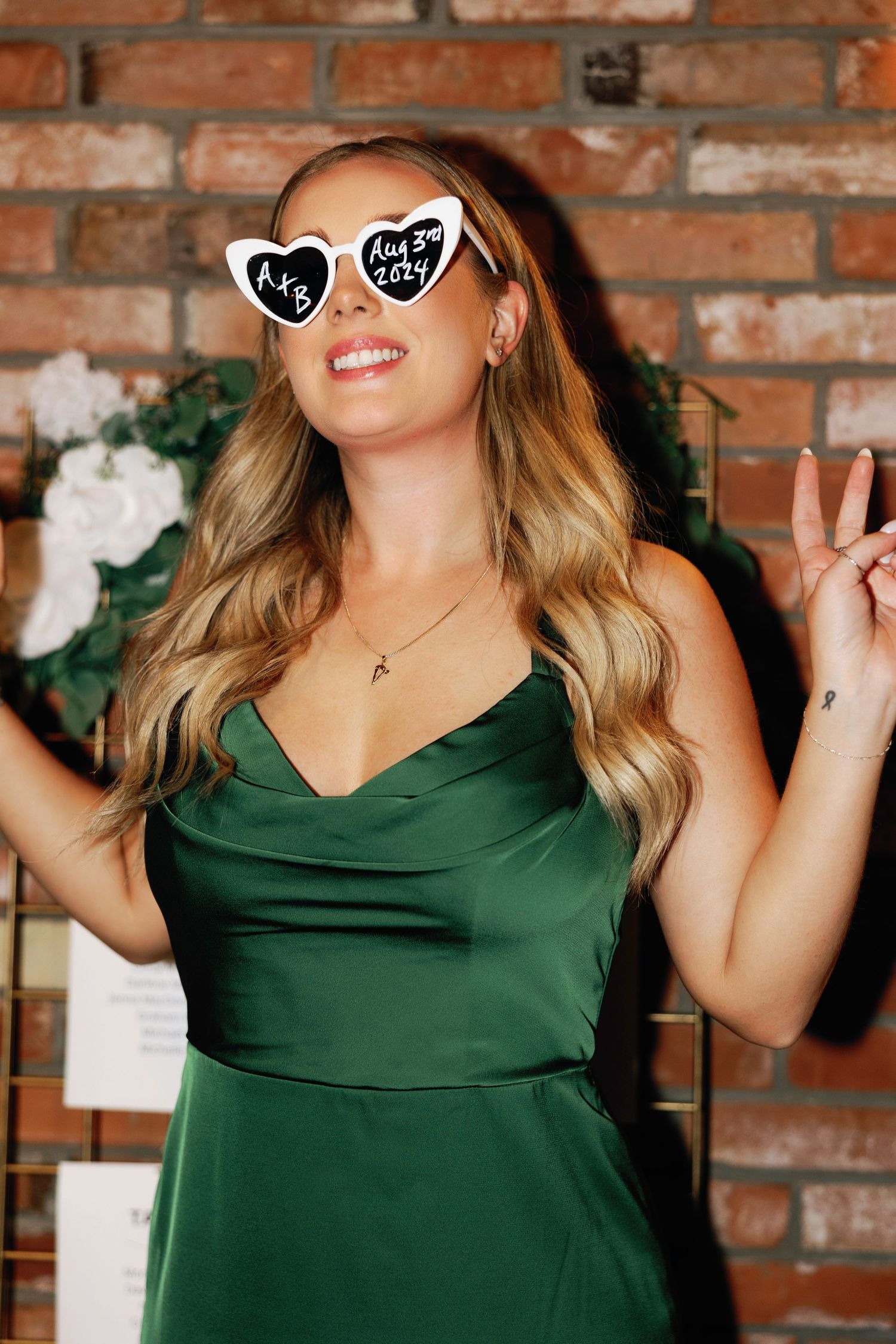 Woman in emerald green dress poses playfully against brick wall wearing heart-shaped sunglasses.
