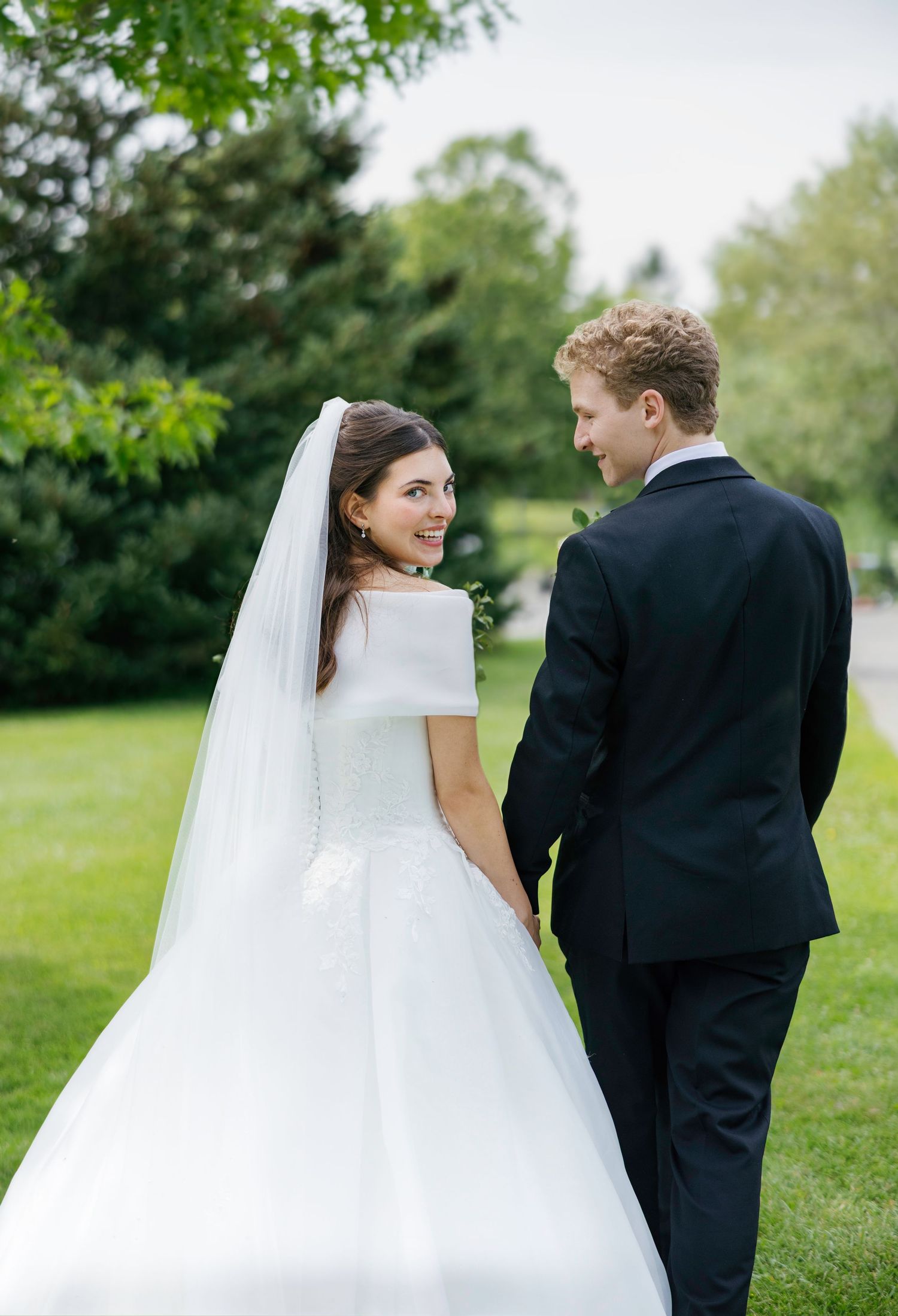 A bride in a white gown and veil holds hands with a groom in a black suit outdoors on a green lawn.
