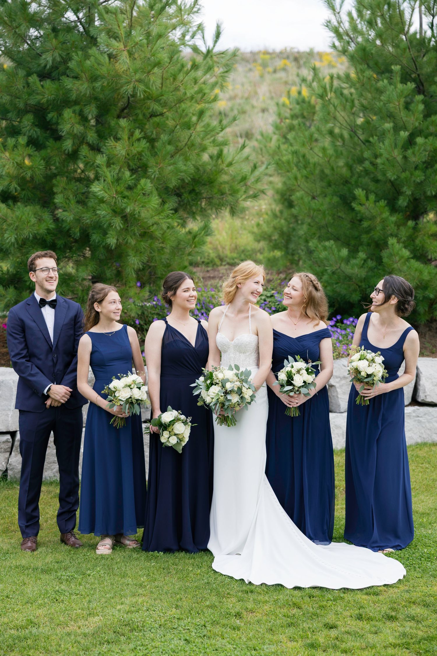 Wedding party in navy blue attire and white dresses stands in line on grassy hill with evergreen trees in background.