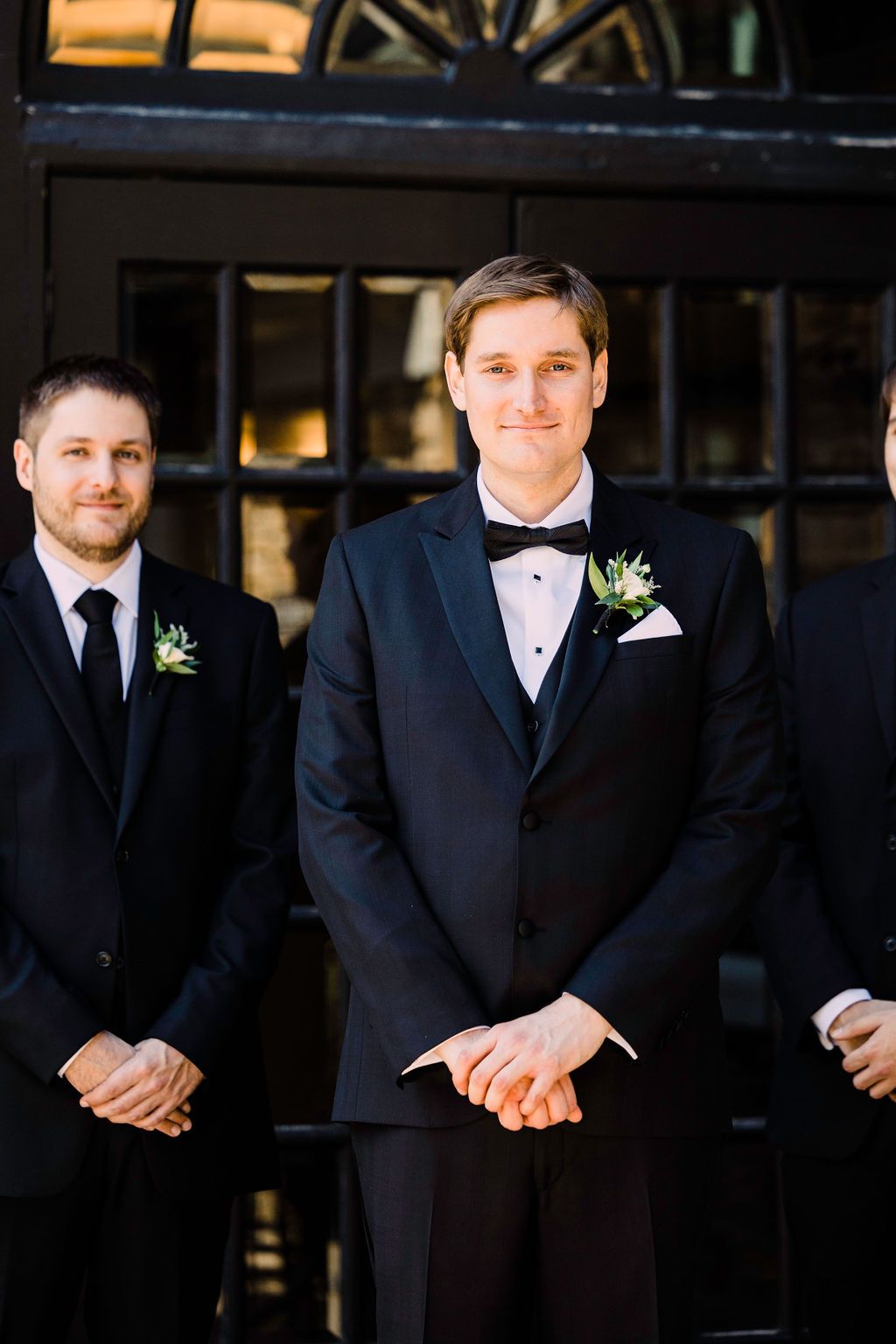 Groomsmen in black tuxedos and bow ties stand together in a formal wedding party lineup.