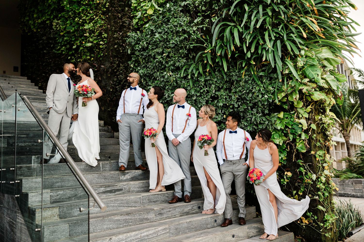 Wedding party in white and gray attire poses on stone steps with lush greenery backdrop.