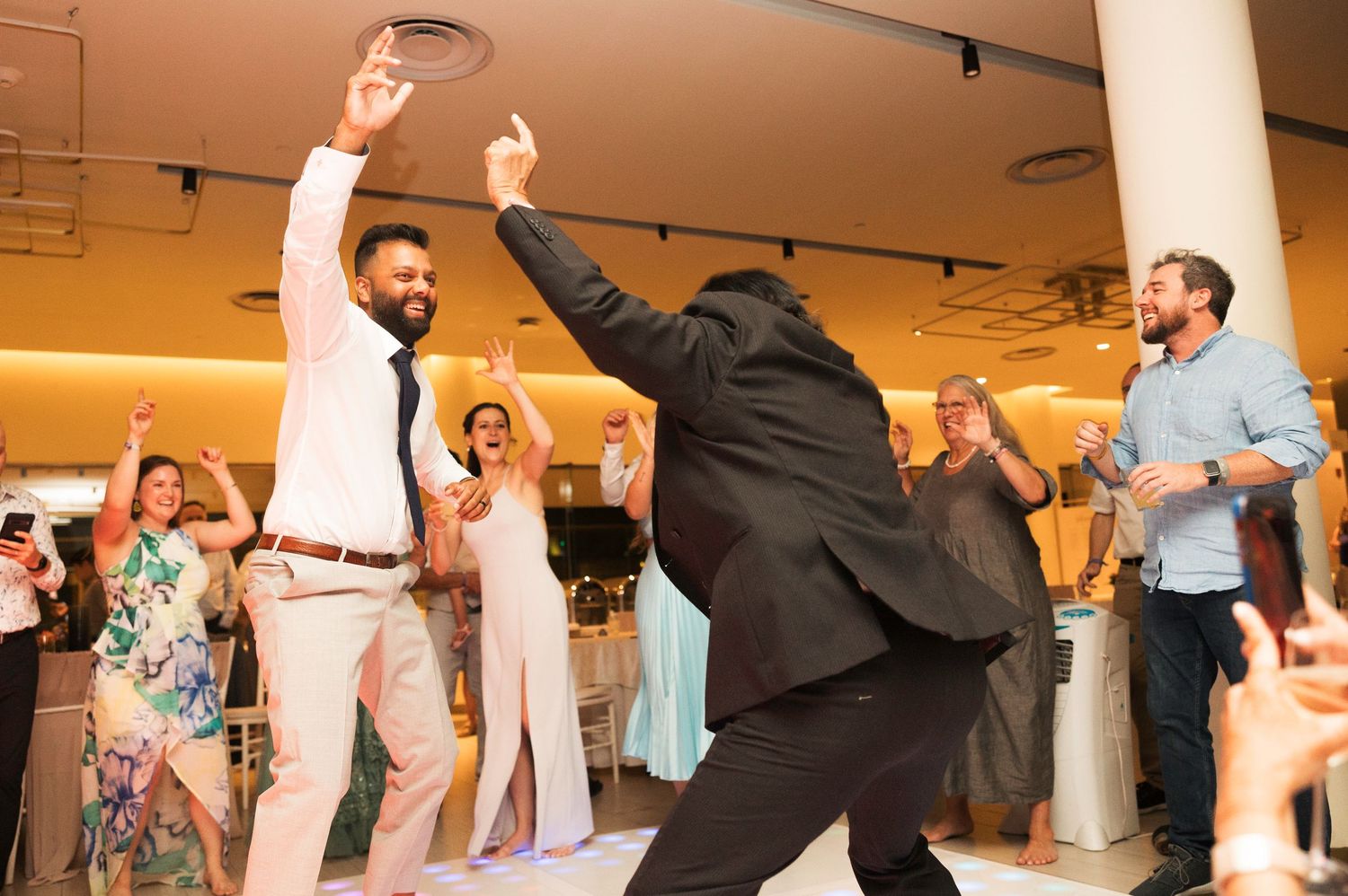 Wedding guests dance and celebrate together on a reception dance floor with festive lighting.