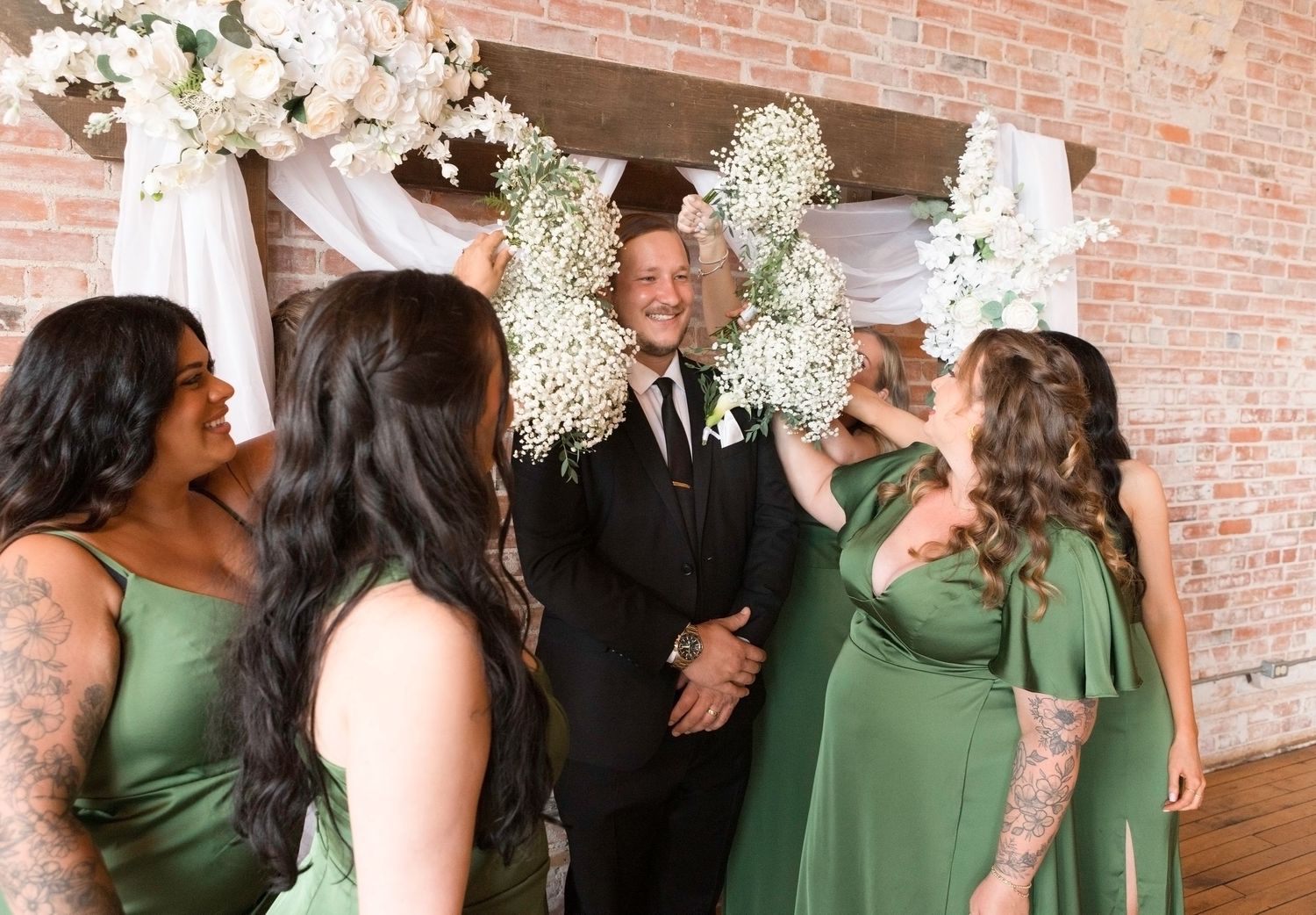 A group of bridesmaids in matching green dresses share a candid laughing moment at a brick-walled venue.