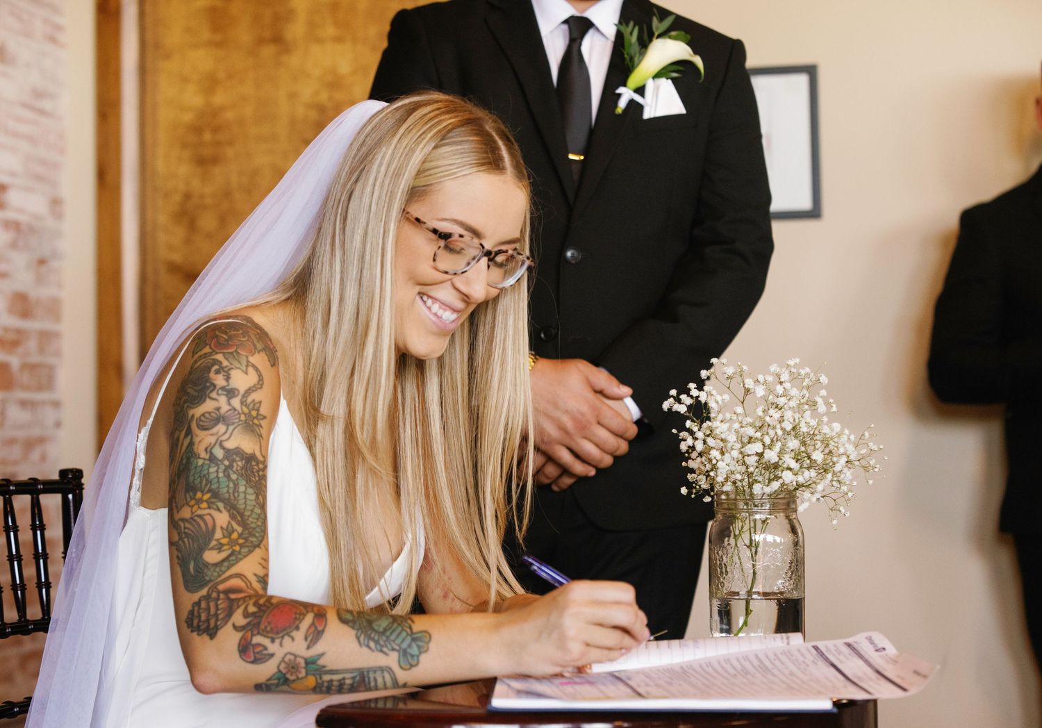 A bride wearing glasses and a white dress with visible arm tattoos signs documents while holding baby's breath flowers.