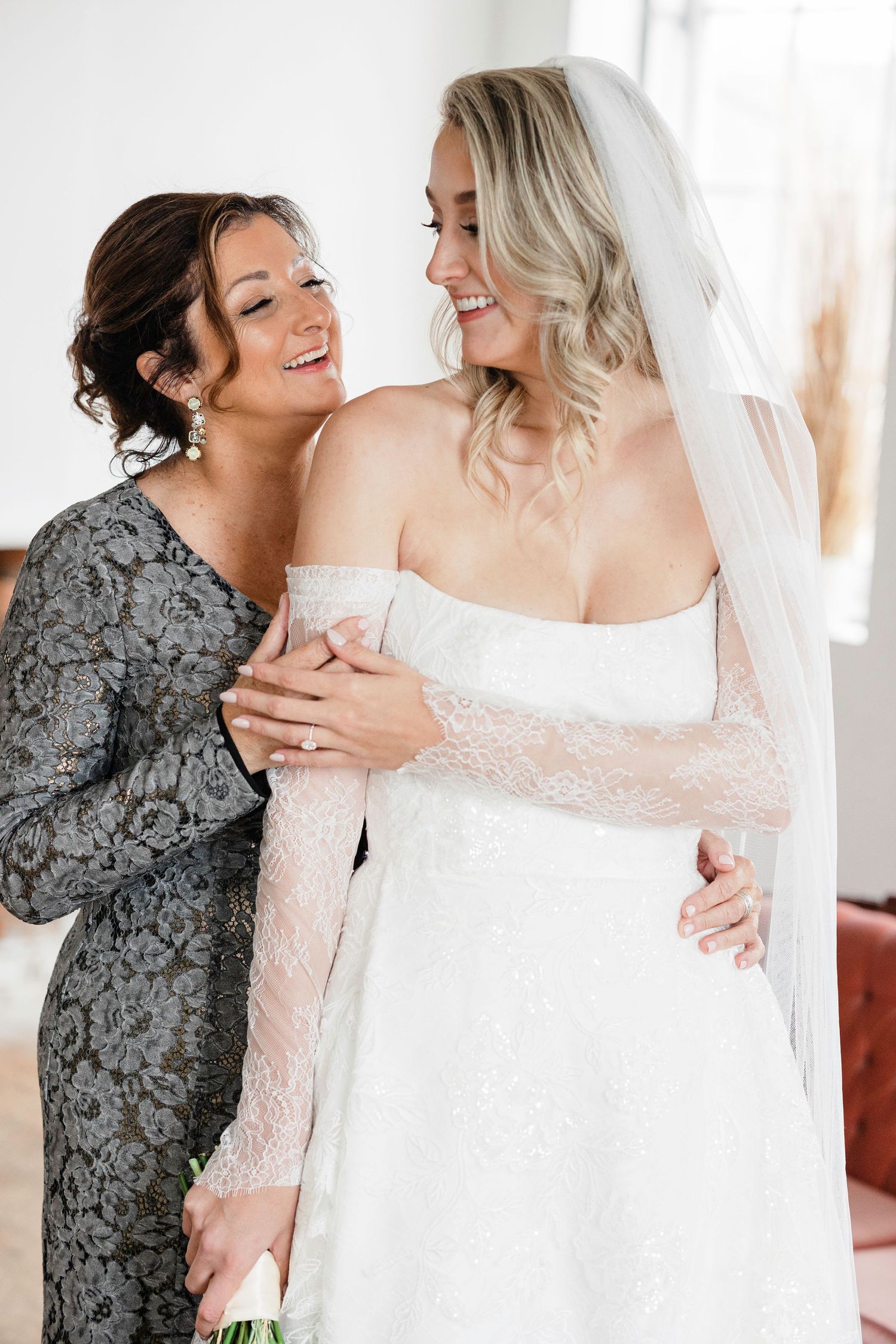 Two women share an emotional embrace during a wedding celebration, one wearing a white gown and the other in grey lace.