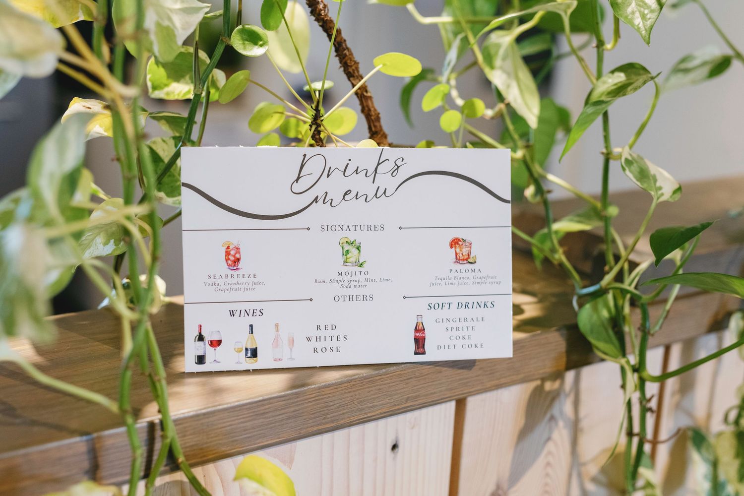 Wooden drink menu sign decorated with green foliage displays beverage options.
