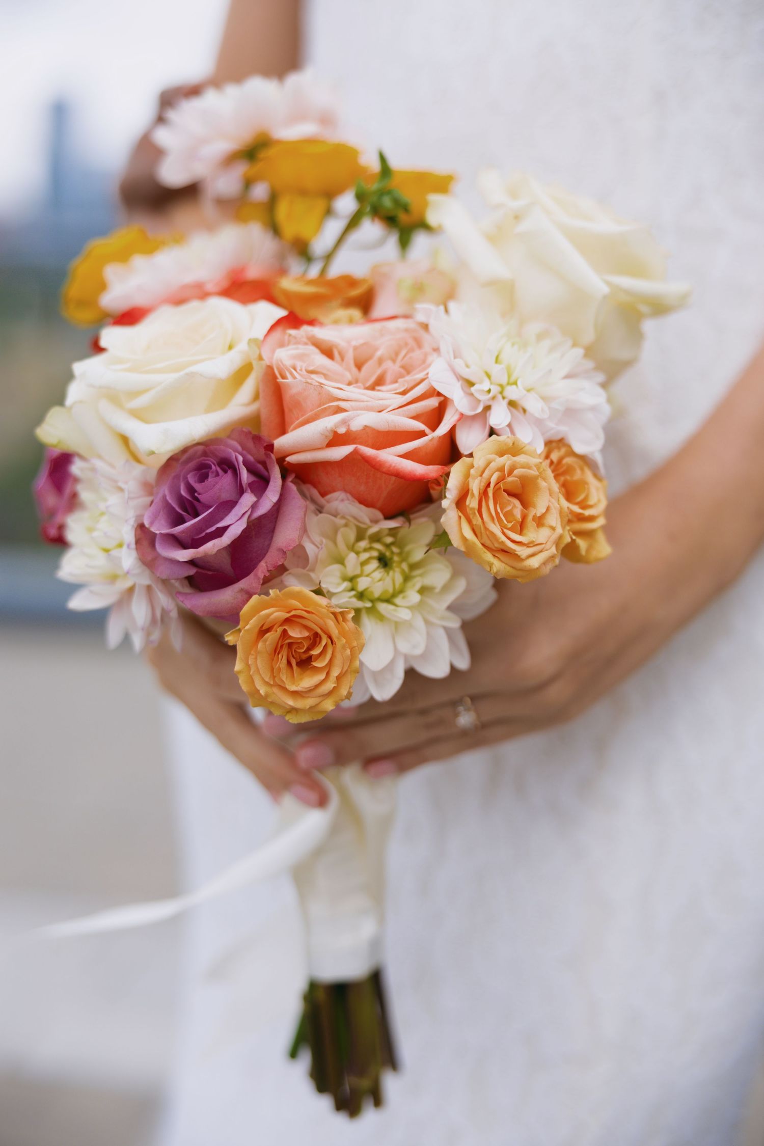 Bride holding a colorful wedding bouquet with peach, purple, orange, and white roses and dahlias wrapped in white ribbon.