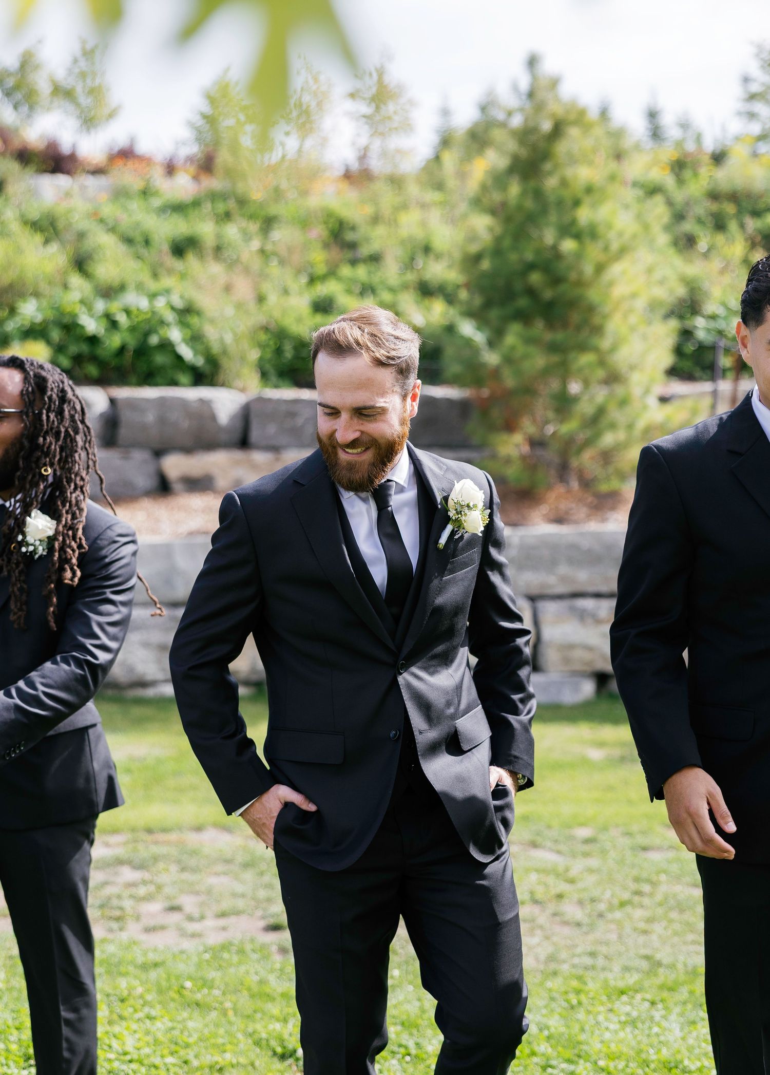 Groomsmen in black suits laugh together while bending down in a candid moment on grass outdoors.